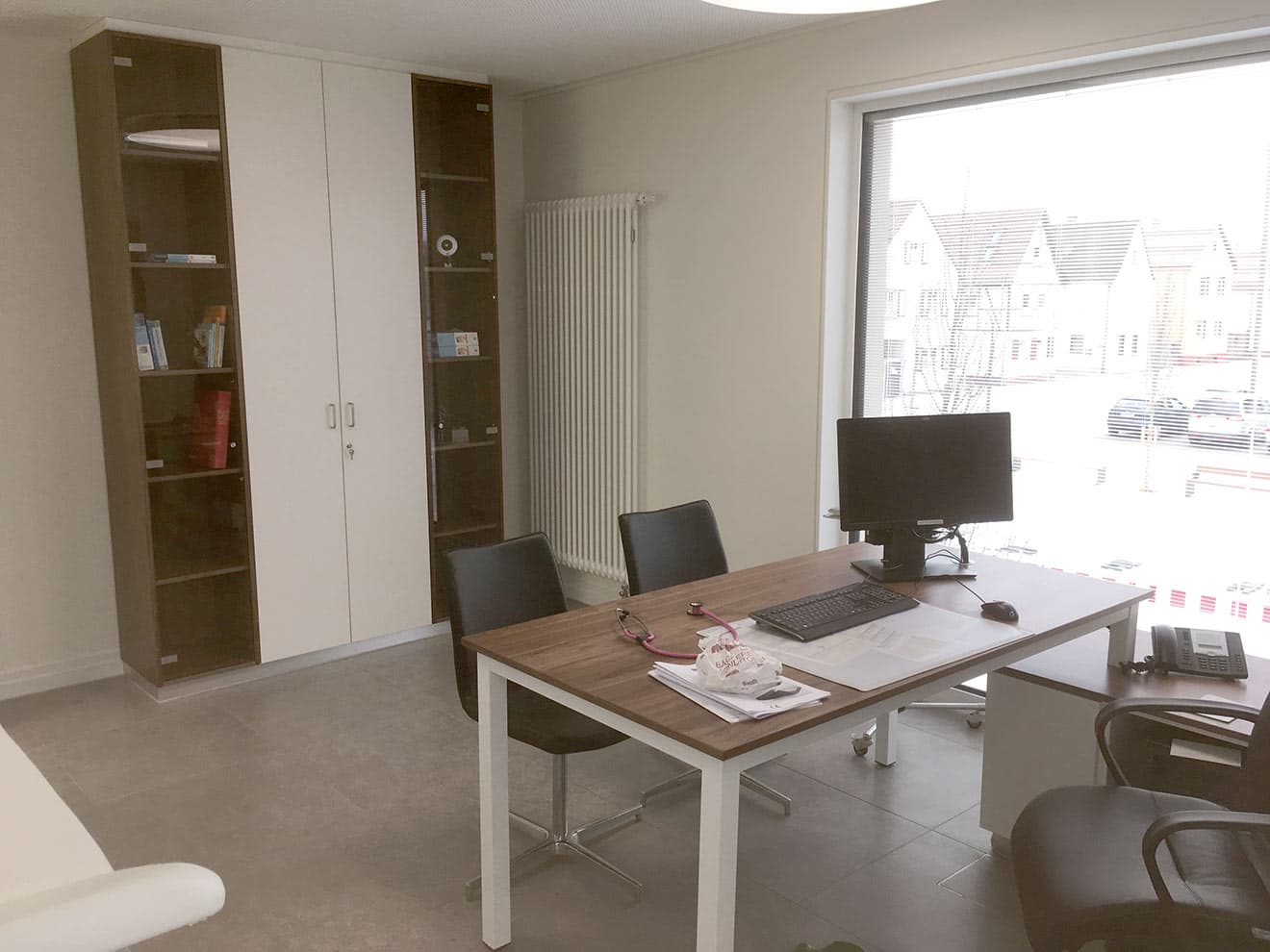 Sprechzimmer der Arztpraxis mit Schreibtisch und raumhohem Schrank mit Glastüren.