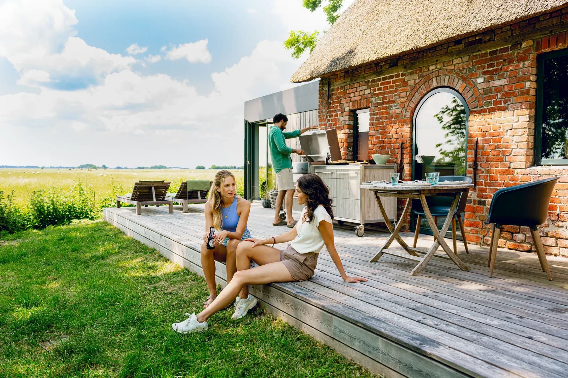 Freunde sitzen auf einer Holzterrasse vor einem reetgedeckten Backsteinhaus, während im Hintergrund jemand an einer BURN-Outdoor-Küche grillt