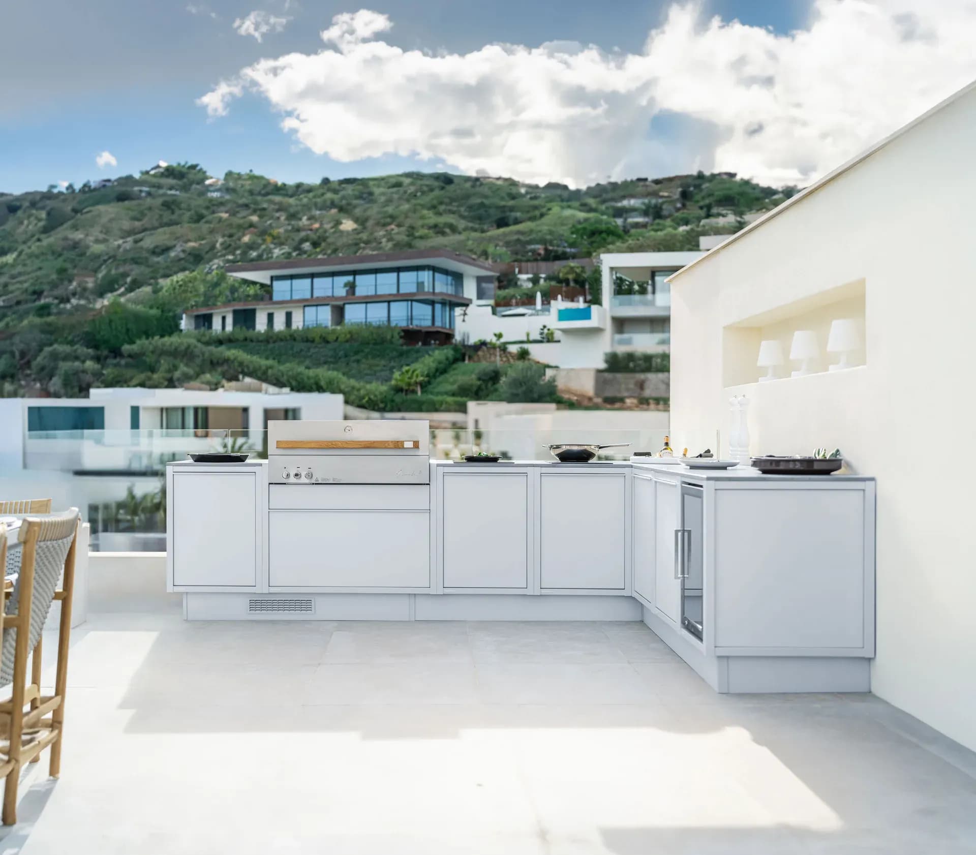 Helle Outdoor-Küche mit Edelstahlgrill auf einer sonnigen Terrasse, dahinter ein Hang mit Villen und das Meer