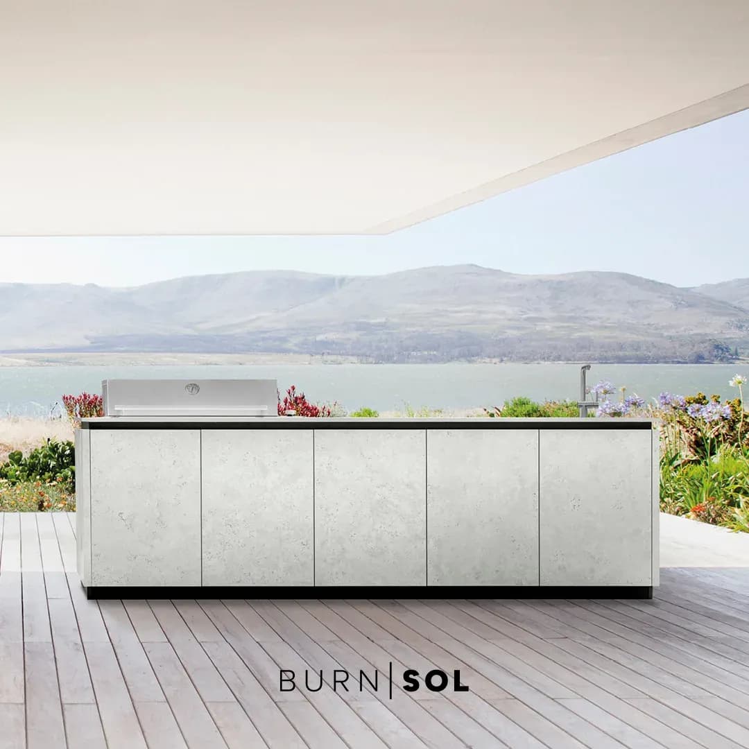 Helle BURN-SOL-Outdoor-Küche mit Keramikfronten und Edelstahlgrill auf einer überdachten Terrasse mit Blick auf einen See und Hügel