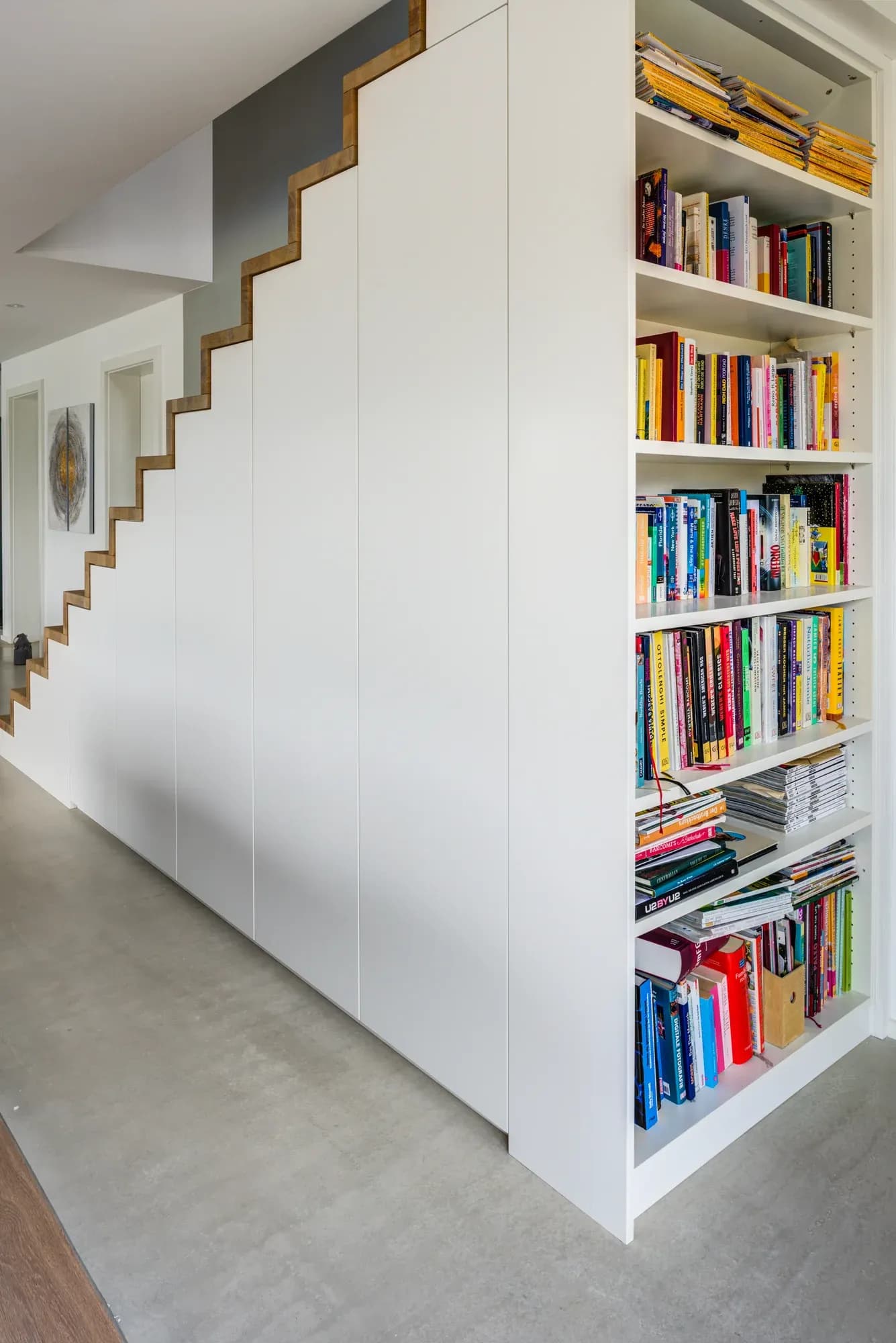 Weiße, grifflose Schrankfronten unter der Treppe, die der Stufenlinie folgen, daneben ein eingebautes Bücherregal.