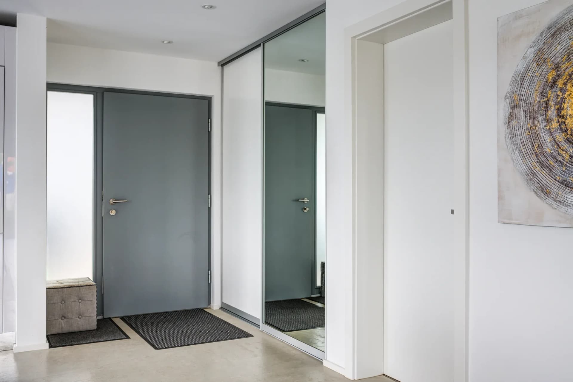 Flur mit grauer Eingangstür und einem Einbauschrank mit Spiegel- und Mattlackfronten, davor ein gepolsterter Hocker und ein rundes Wandbild.
