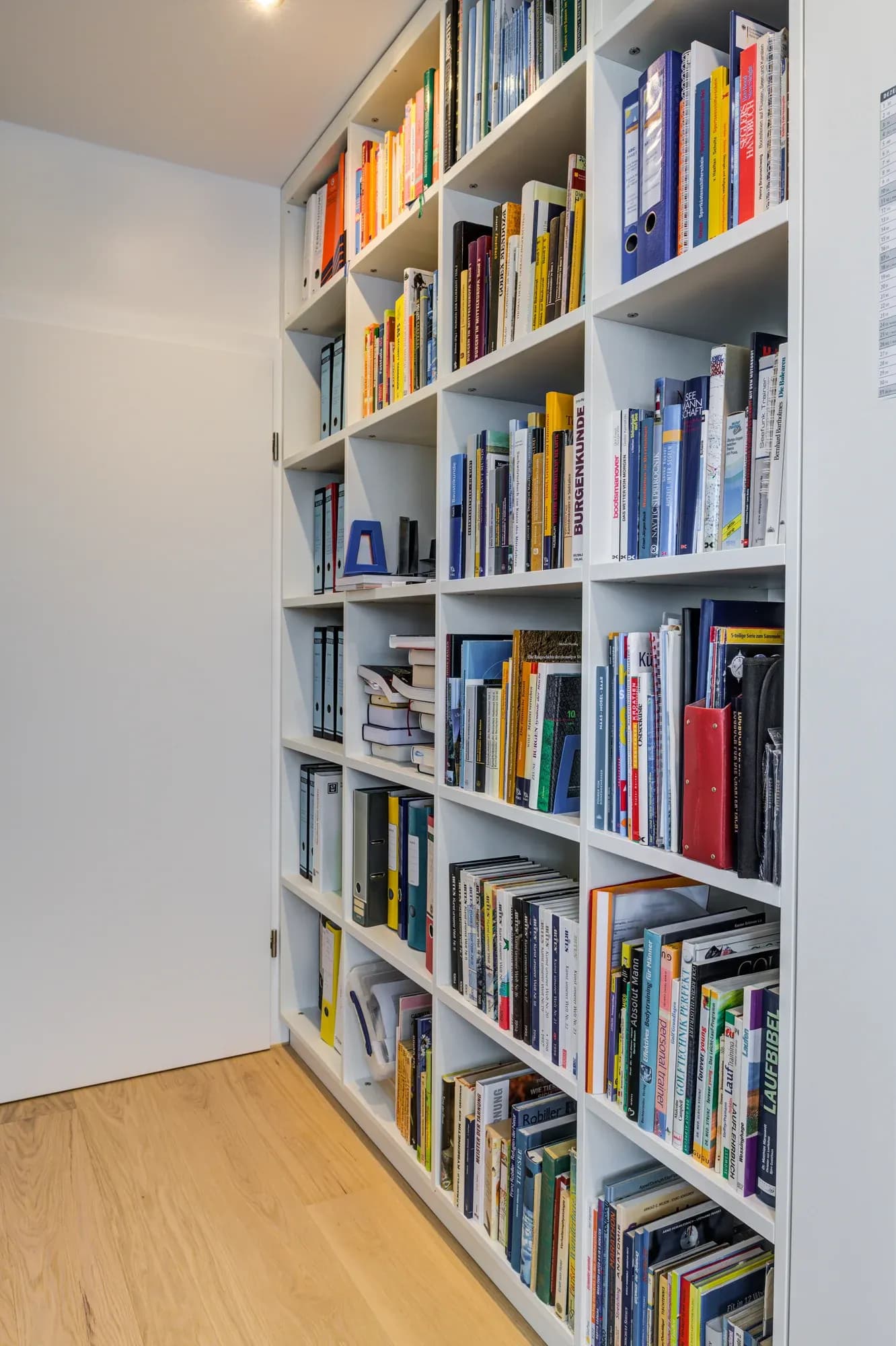 Raumhohe weiße Bücherwand über Eichenparkett, gefüllt mit farbig sortierten Büchern