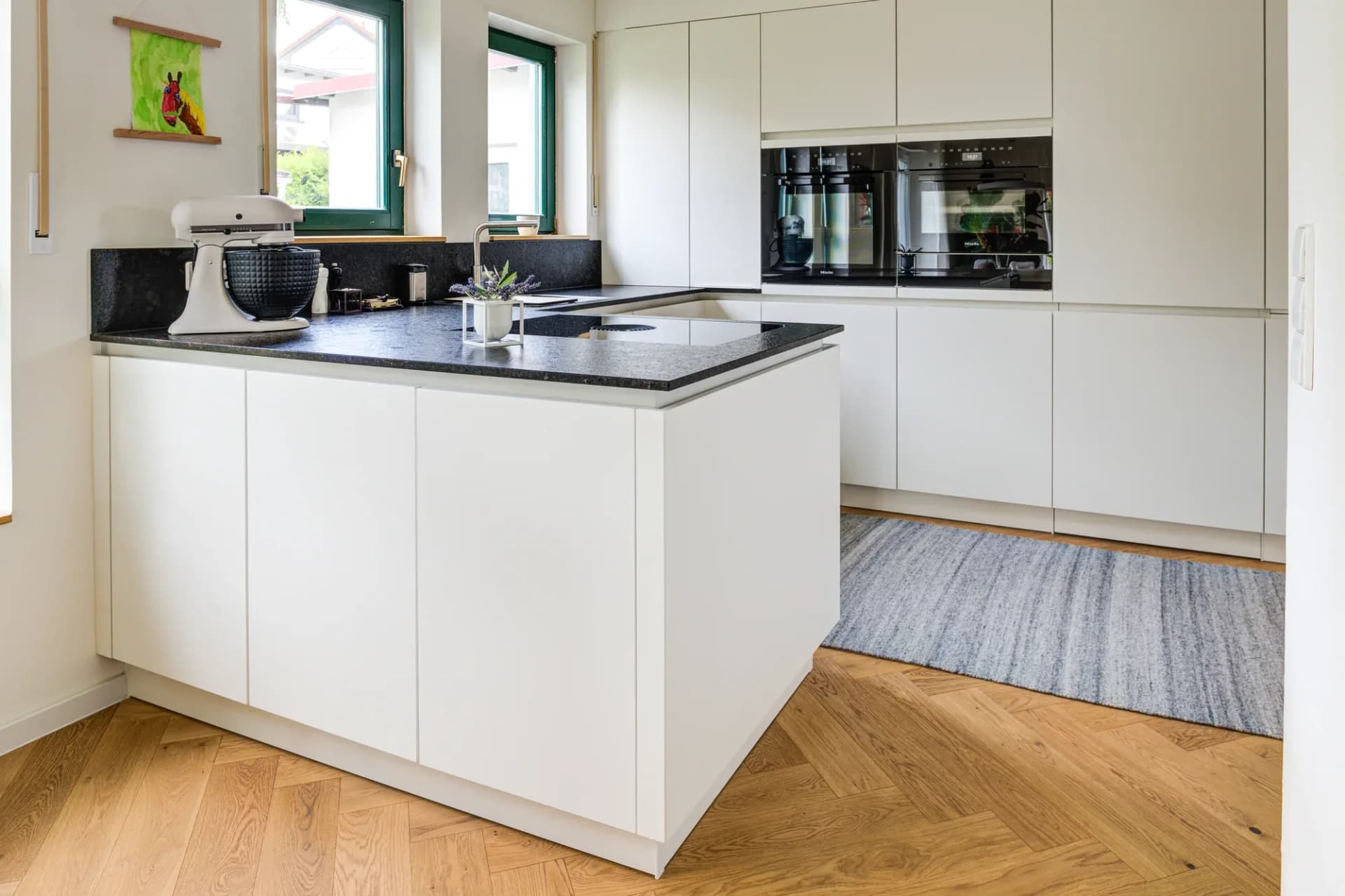 Blick aus der Tür in die weiße U-Küche mit dunklem Natursteinabschluss, grünen Holzfenstern und Eiche-Fischgrätparkett.