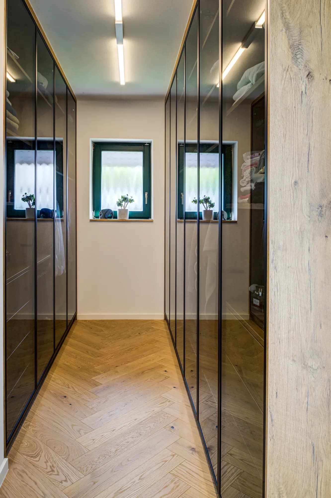 Begehbarer Schrank mit raumhohen Fronten aus bronzefarbenem Glas beidseits eines Fensters, darunter Eiche-Fischgrätparkett.
