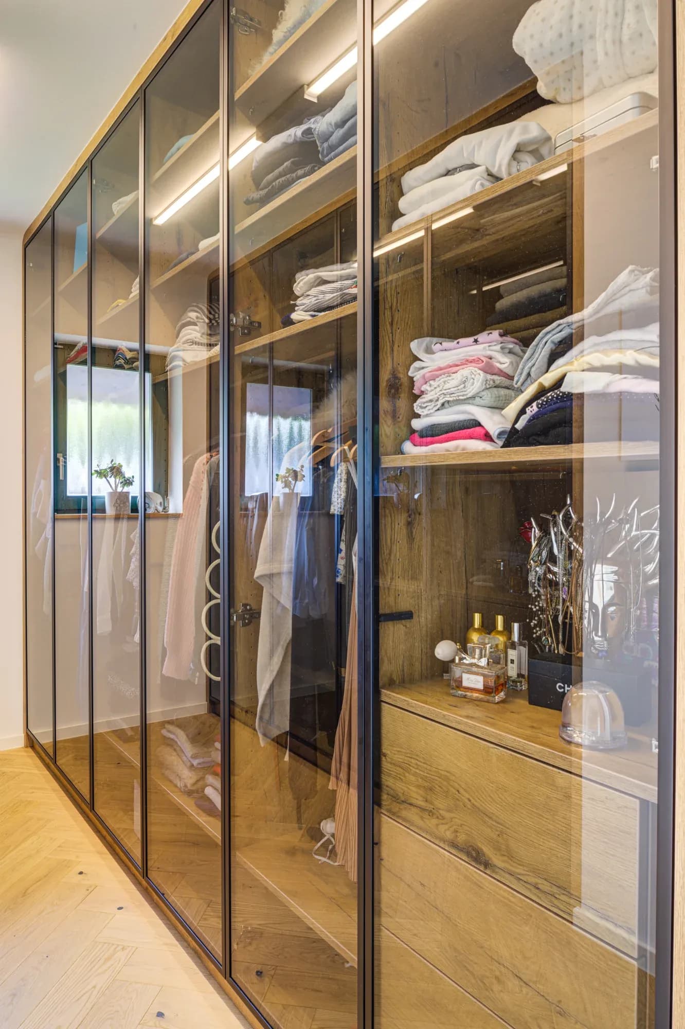 Blick in den begehbaren Schrank: bronzefarbene Glasfronten über eichefurniertem Innenausbau mit beleuchteten Fächern und Kleiderstangen.