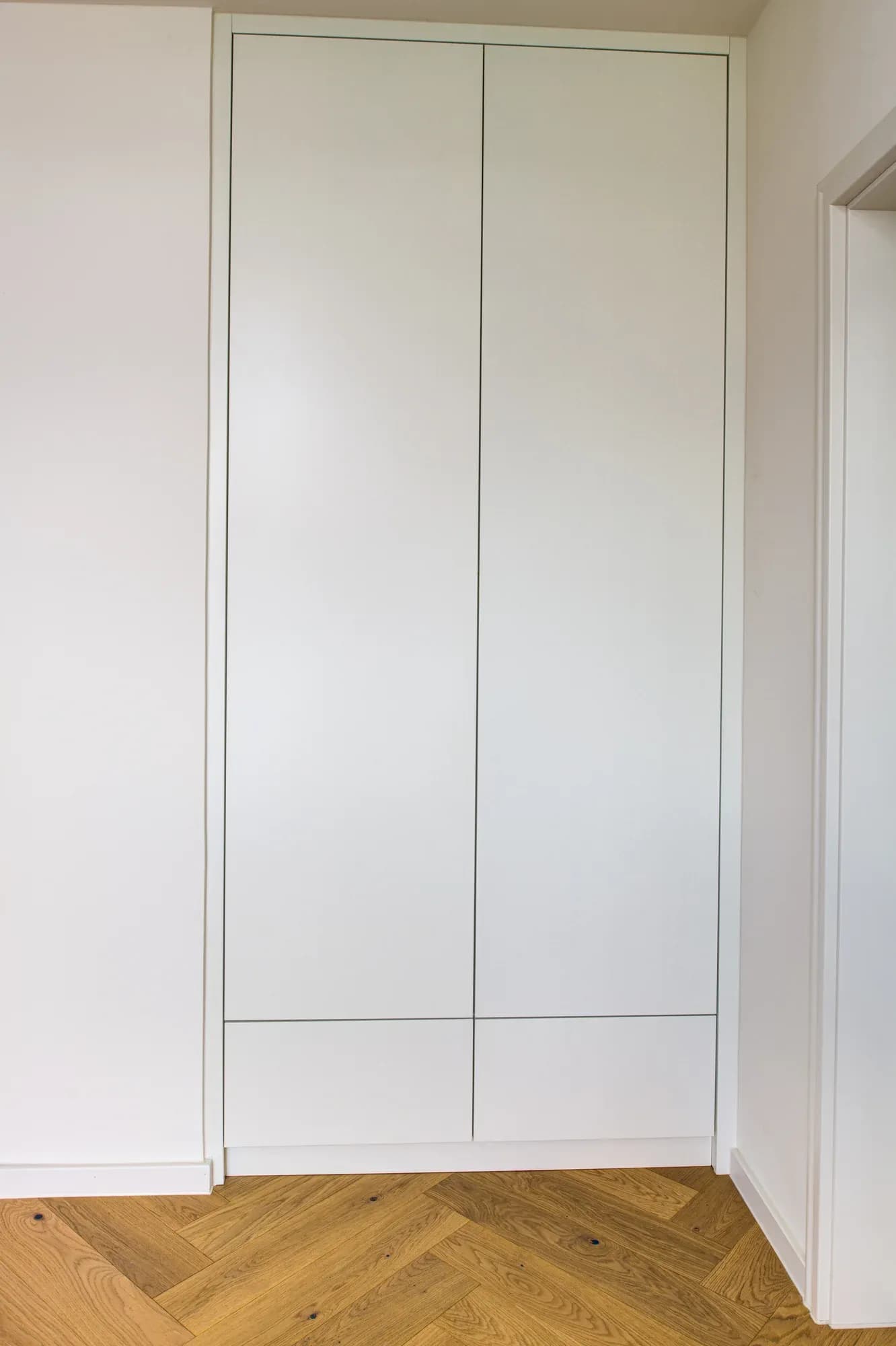 Raumhoher weißer Einbauschrank mit grifflosen Fronten in einer Nische über Eiche-Fischgrätparkett.