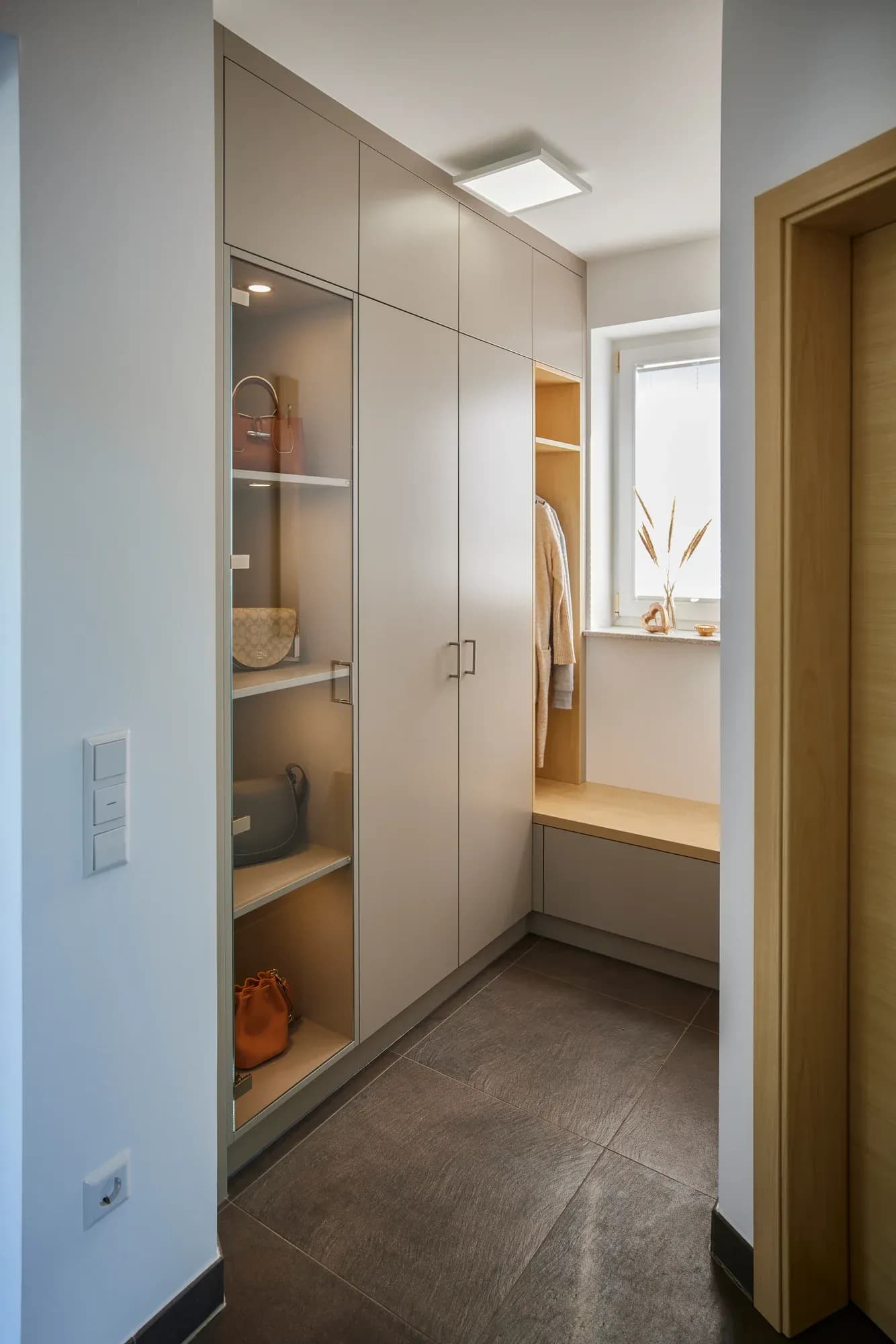 Schmaler Flur mit raumhohem Einbauschrank in Lichtgrau, beleuchteter offener Vitrine, einer Sitzbank aus Holz unter dem Fenster und heller Holz-Zimmertür rechts.