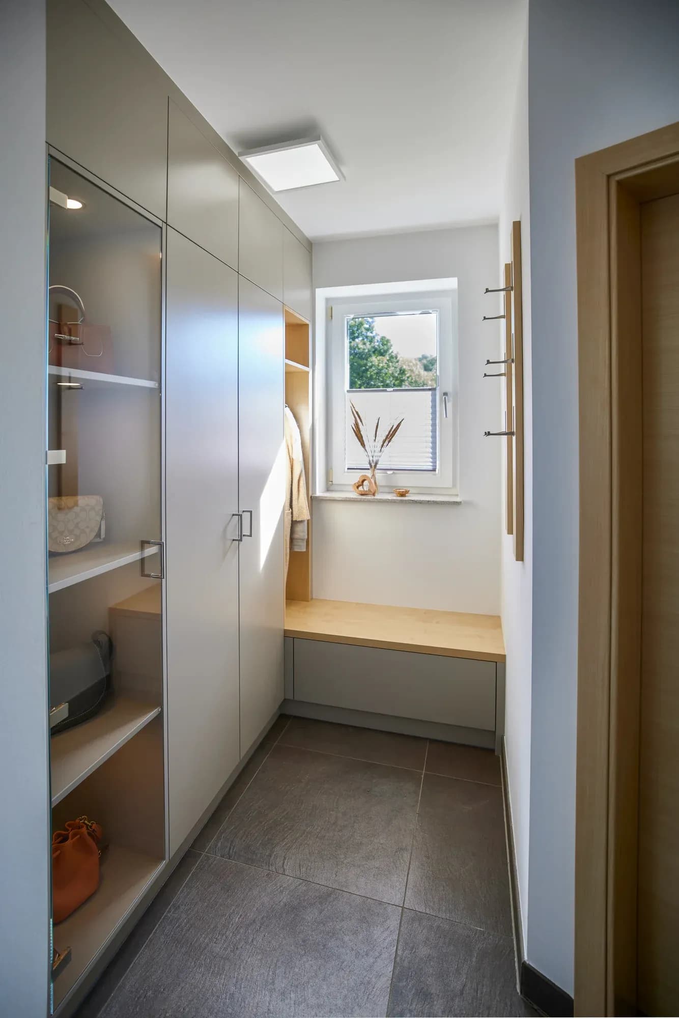 Blick durch den Flur: Garderobenschrank mit gläserner Vitrinentür links, Sitzbank unter dem Fenster und hölzerne Hakenleisten an der Wand rechts.