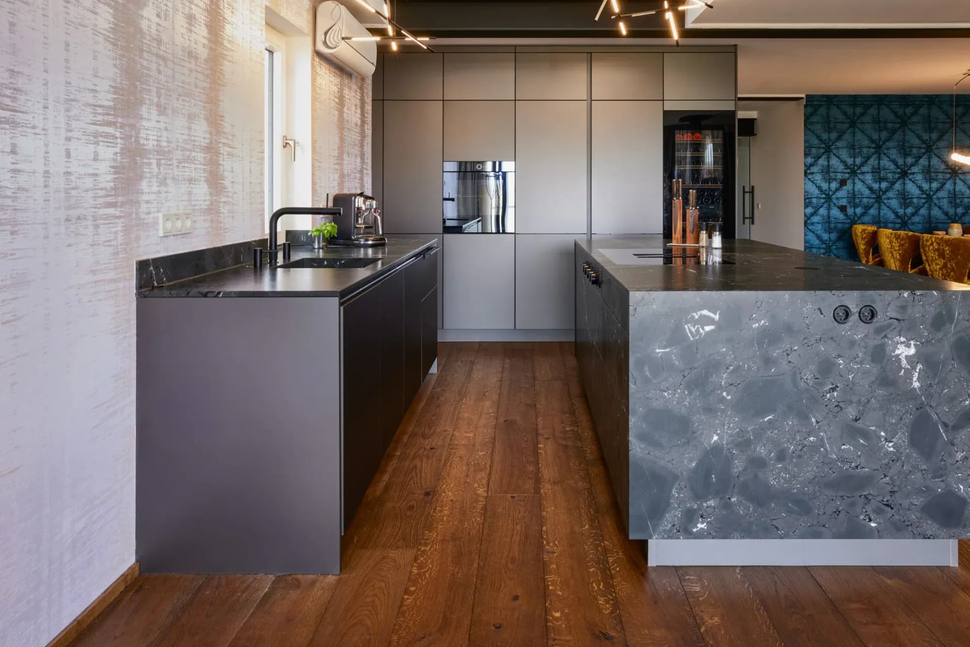 Arbeitsgang zwischen der mattschwarzen Küchenzeile mit Spüle und der Natursteininsel, im Hintergrund die taupefarbene Hochschrankwand mit Backofen und Weinklimaschrank.