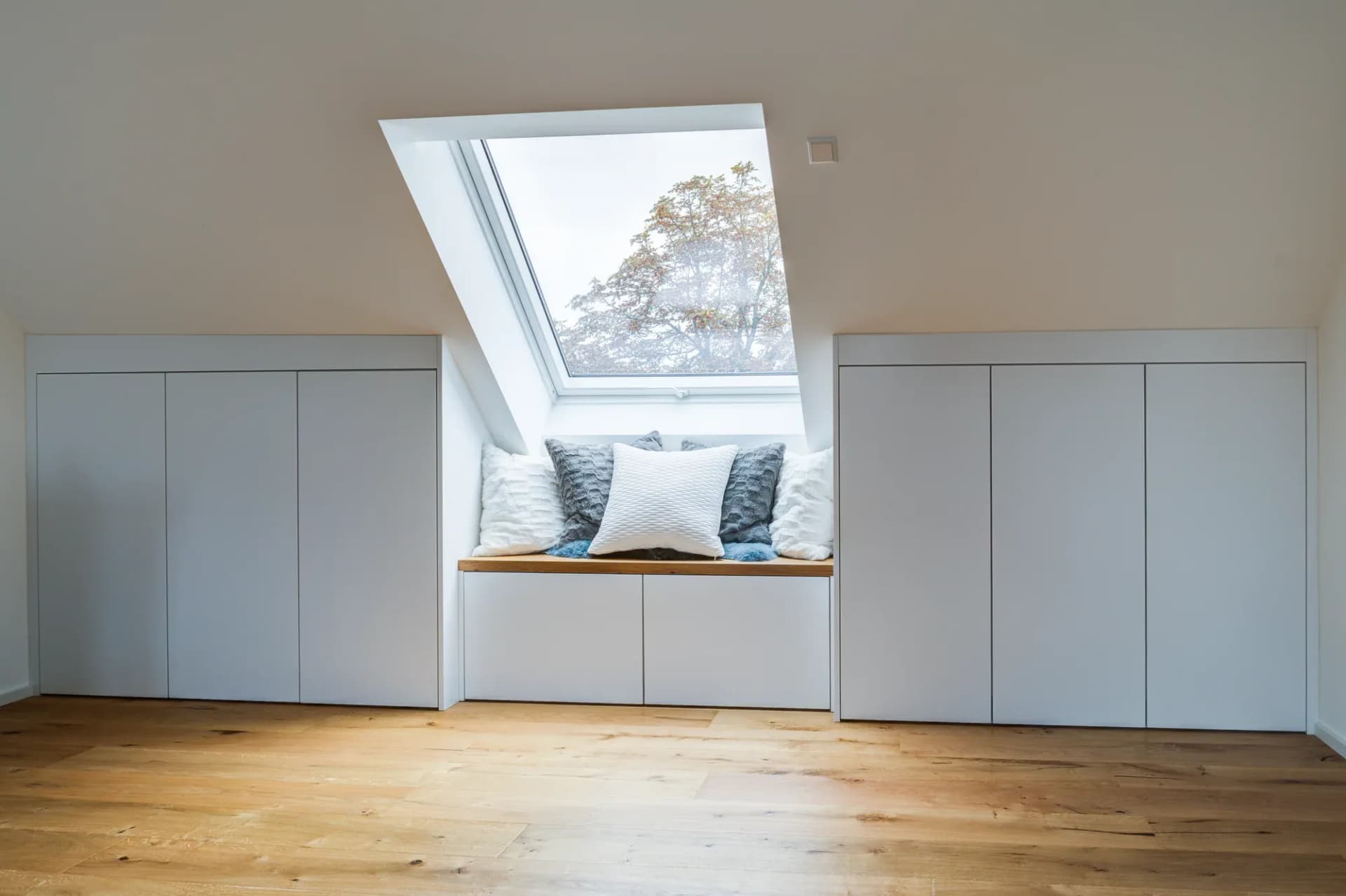 Sitznische aus Eiche unter einem Dachfenster, flankiert von weißen Einbauschränken, im Fenster ein Baum.