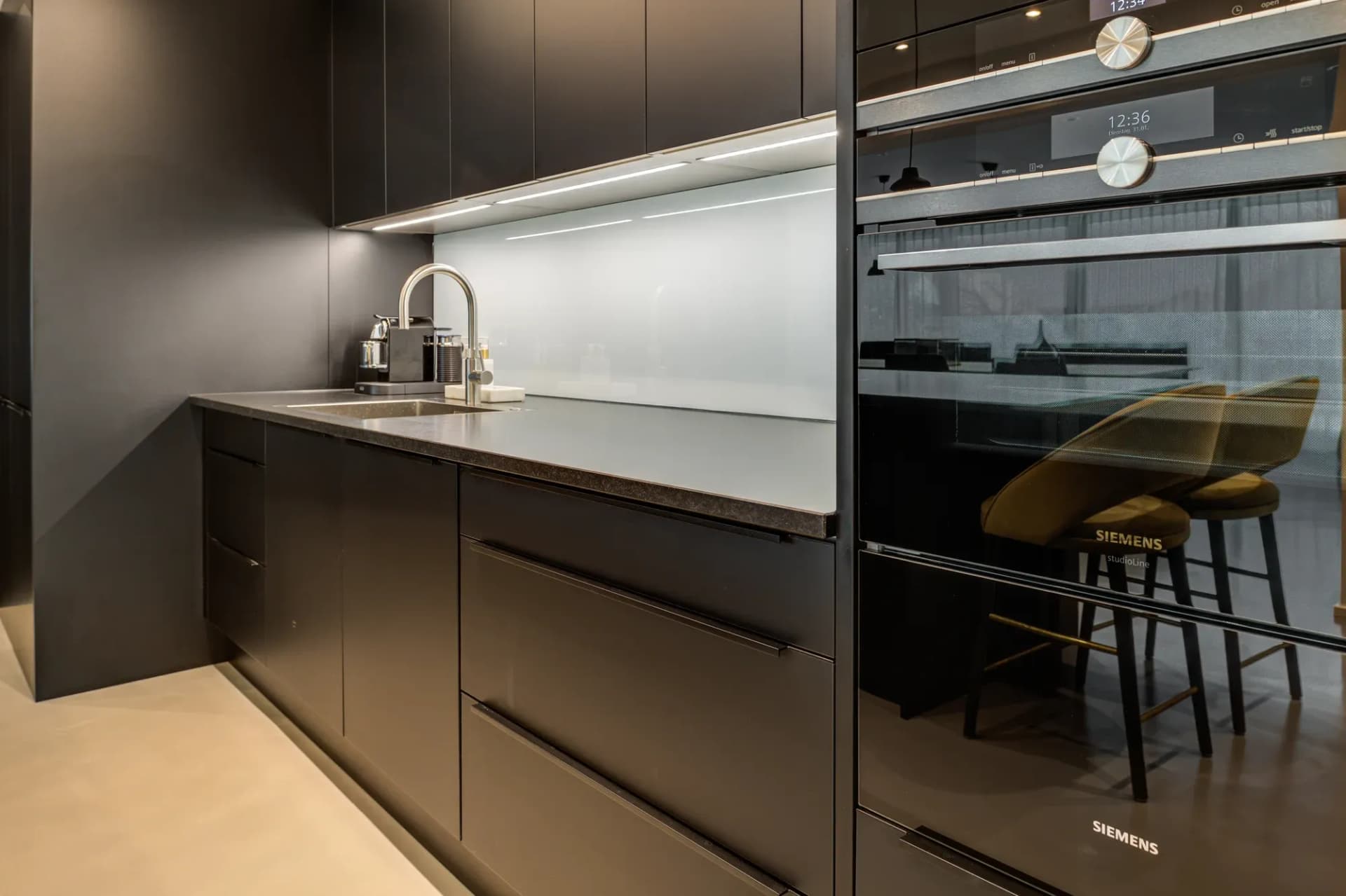Schwarze Küchenzeile mit Glasrückwand, Unterbauleuchte und zwei Siemens-Backöfen.