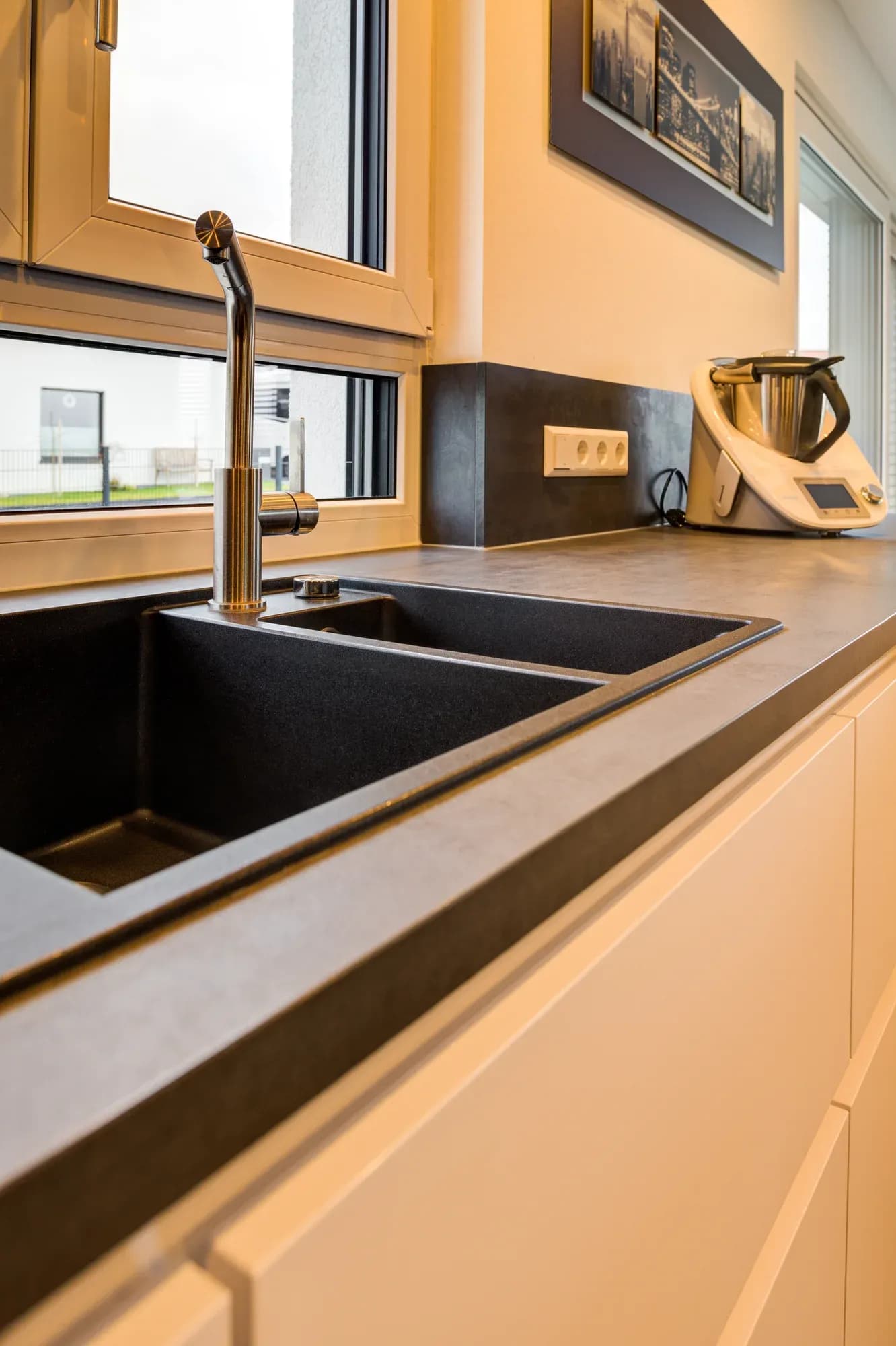 Schwarze Granit-Verbundspüle mit ausziehbarer Armatur im Bronzeton vor dunkler Keramik-Rückwand und Fenster.