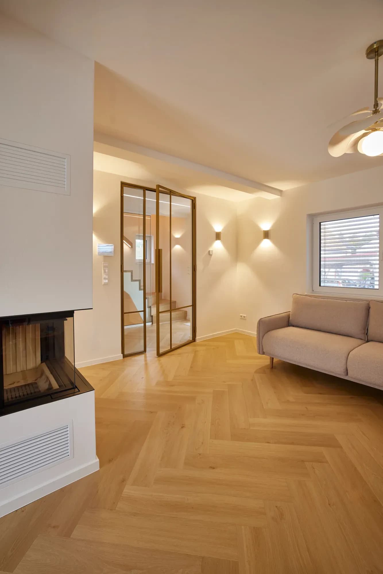 Wohnraum mit weißem Kamin, messingfarben gerahmter Glastür zum Treppenhaus und heller Polstercouch auf Fischgrätparkett.