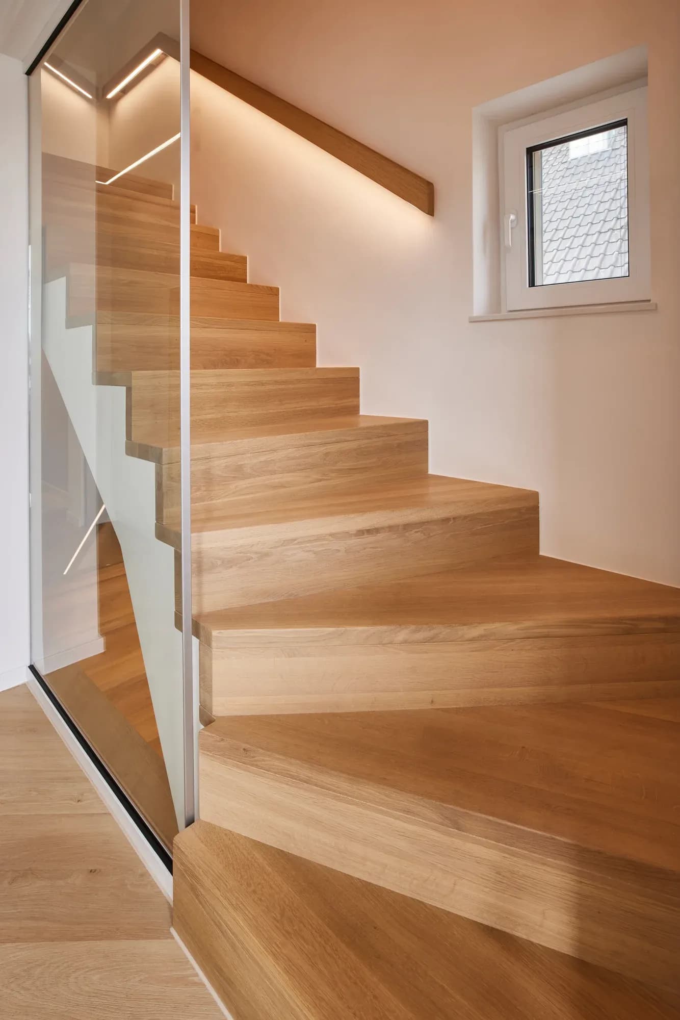 Treppe mit massiven Eichenstufen, Glaswand als Absturzsicherung und einem Eichen-Wandhandlauf mit LED-Beleuchtung.
