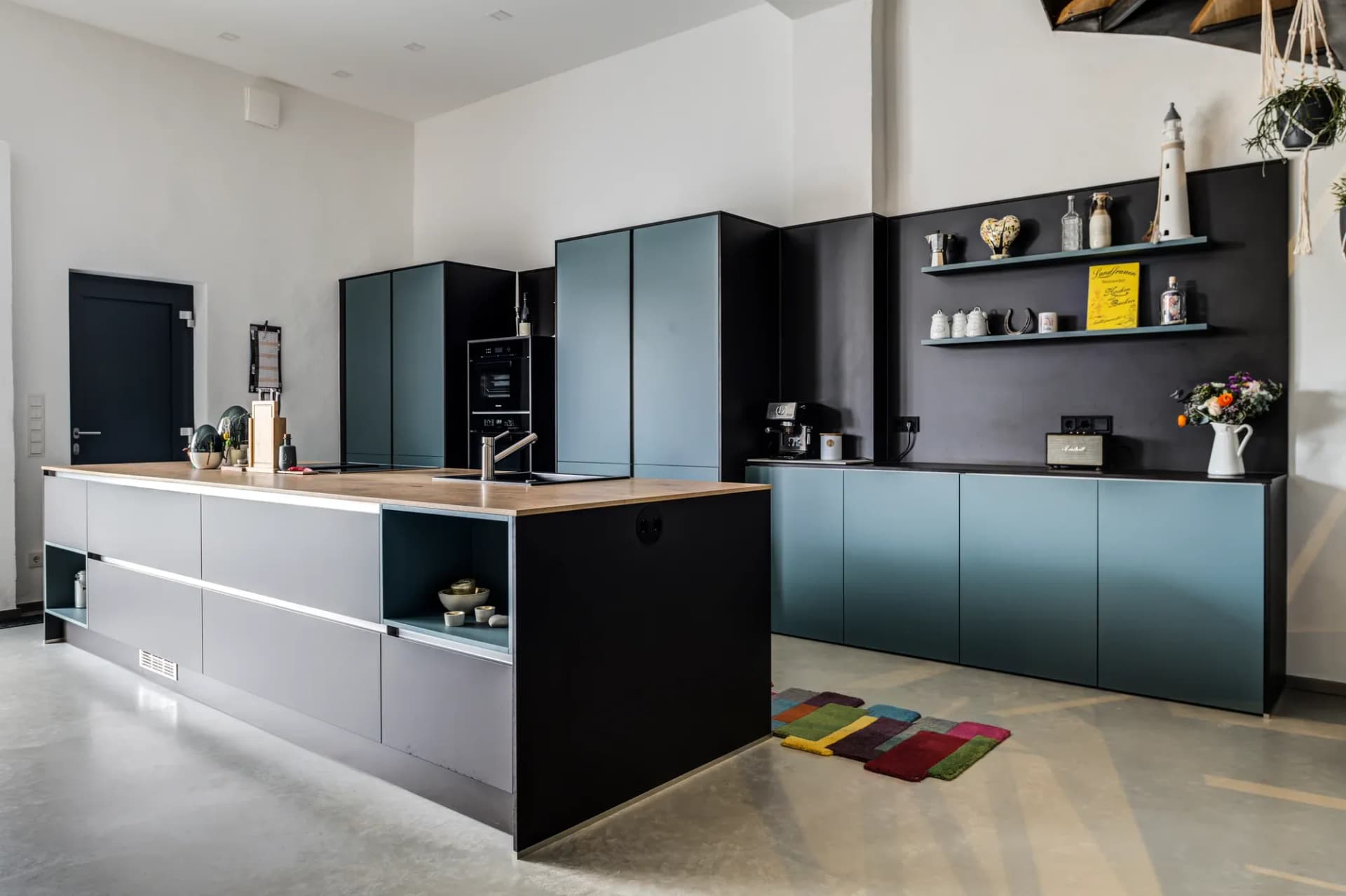 Mattschwarze Gerätewand mit eingebauten Backöfen, davor die Kücheninsel mit Eiche-Arbeitsplatte und Pendelleuchten.