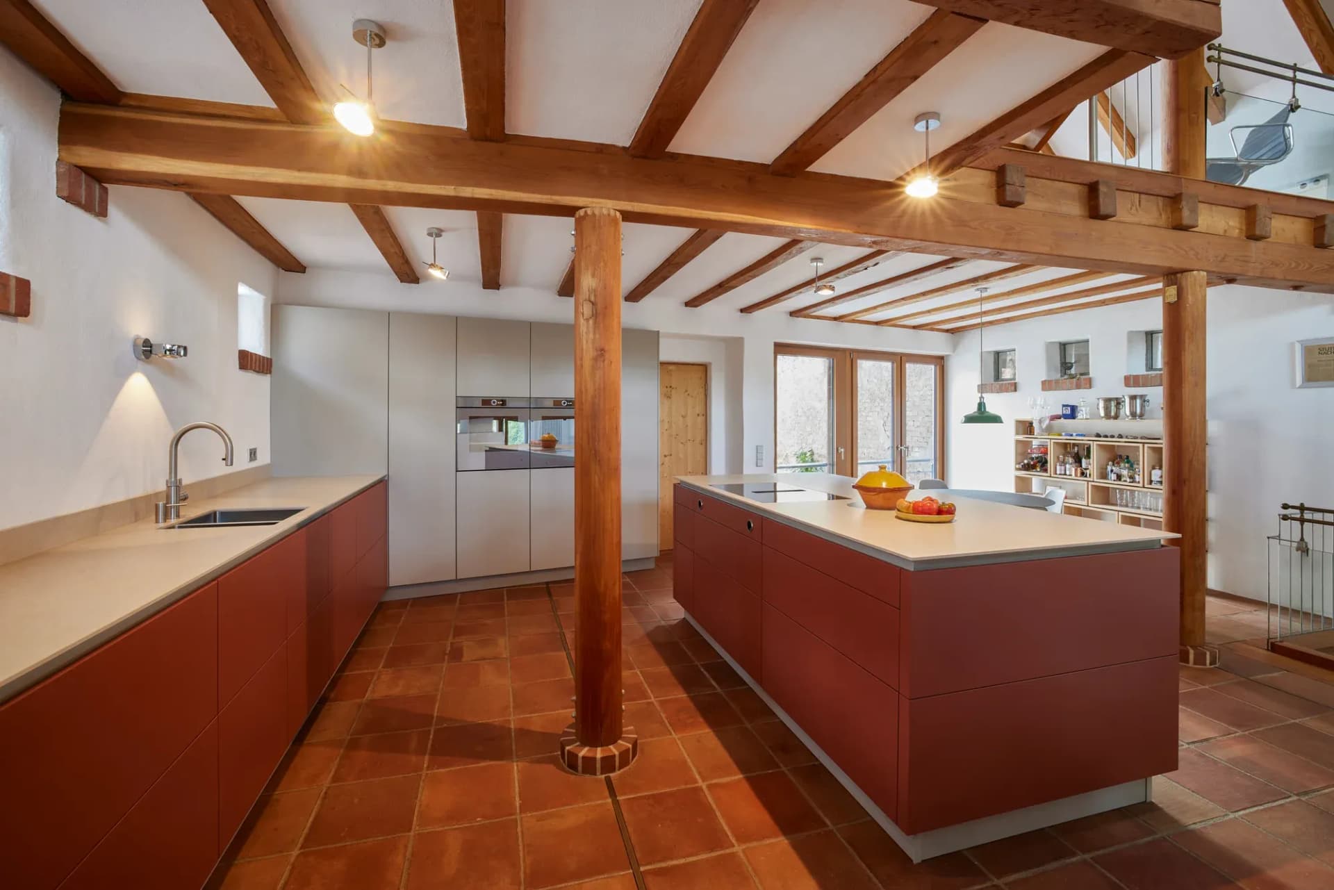 Blick durch den Raum: rote Unterschrankzeile mit Spüle links, tragende Holzstütze in der Mitte und die Kochinsel rechts, im Hintergrund offene Regale am Gartenfenster.