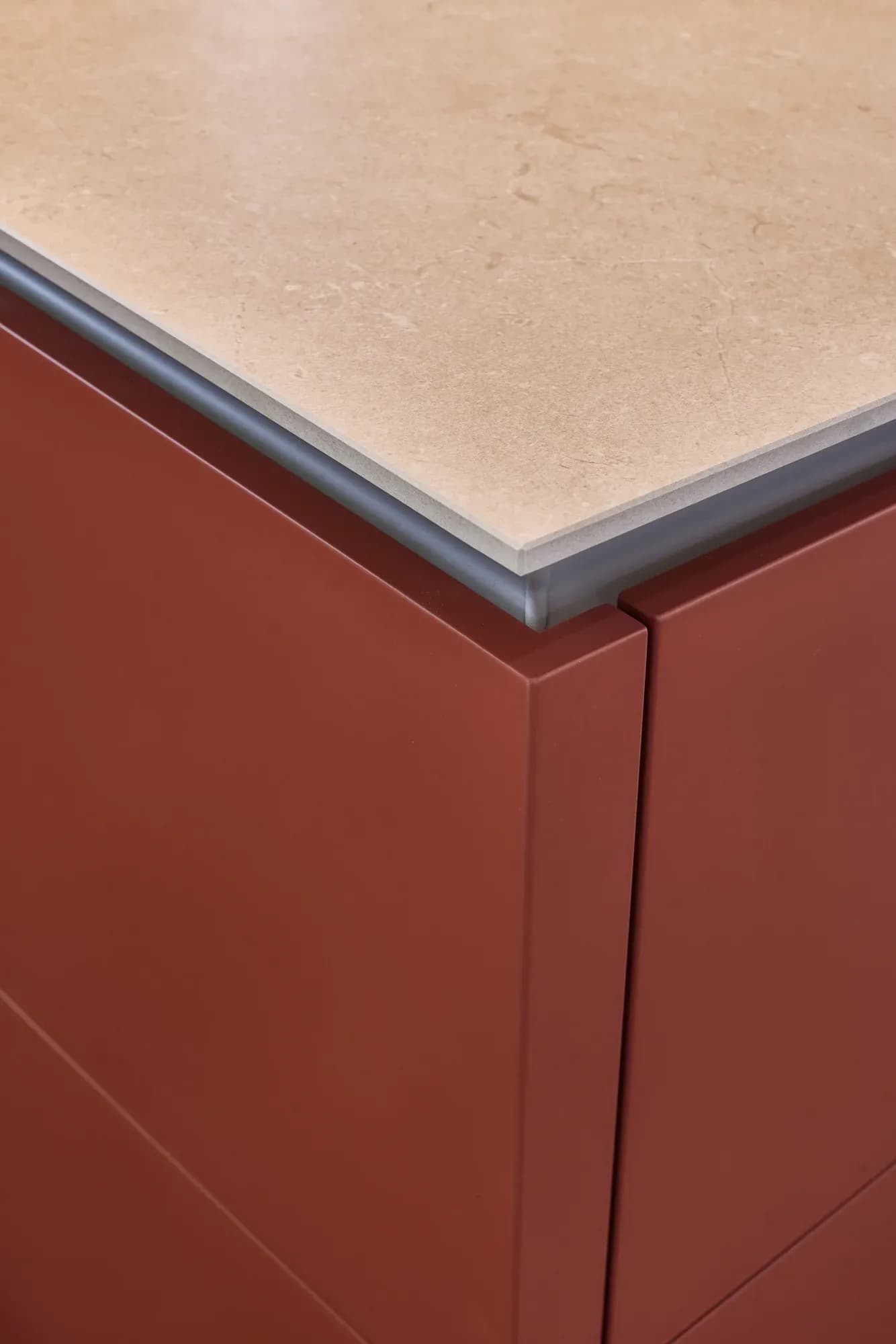 Materialdetail: helle Natursteinplatte auf terrakottaroten Mattlack-Fronten, getrennt durch eine schmale anthrazitfarbene Schattenfuge.