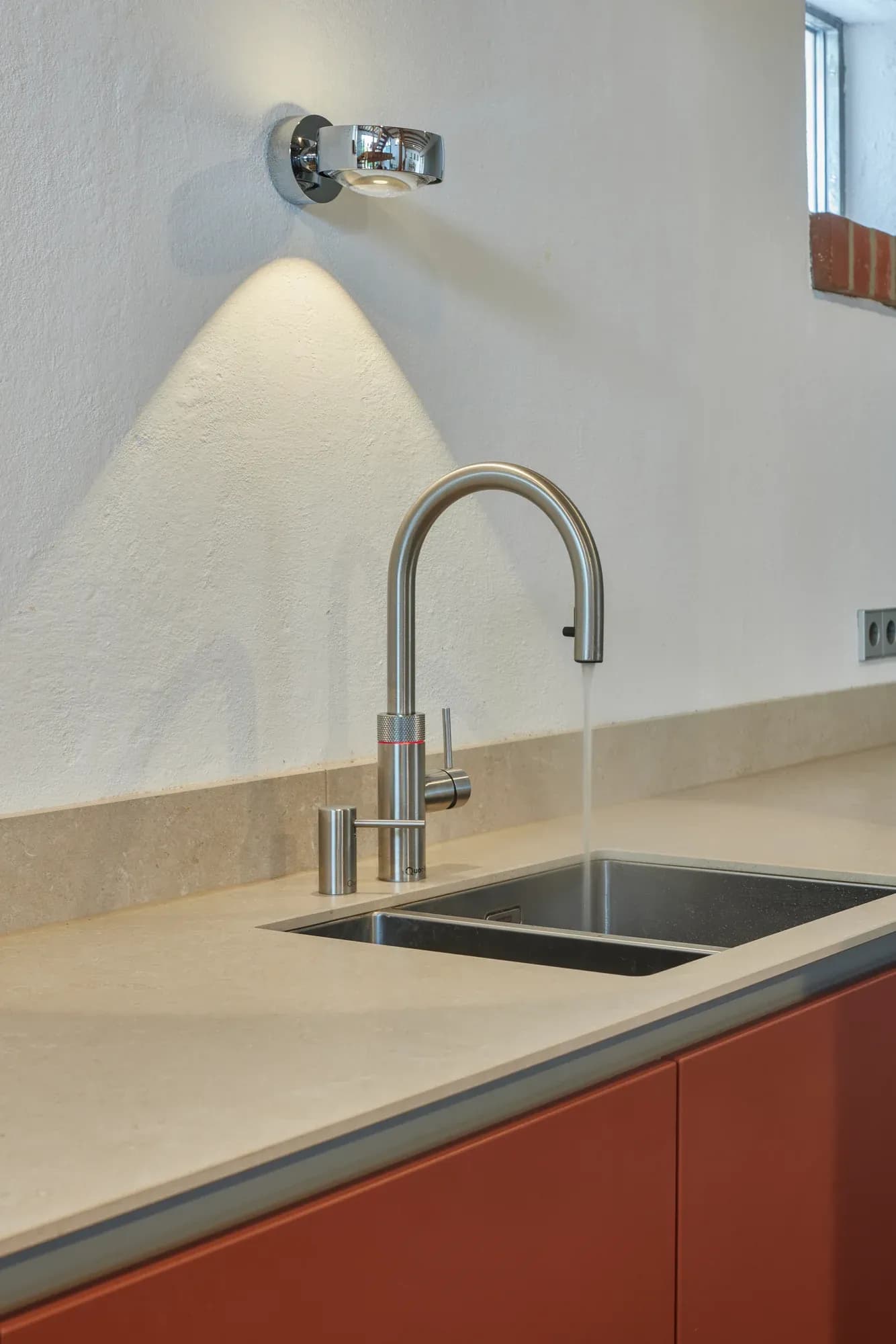 Detail der Spüle: Edelstahl-Armatur mit Kochendwasser-Funktion, aus der Wasser in das Edelstahlbecken läuft, darüber eine verchromte Wandleuchte.