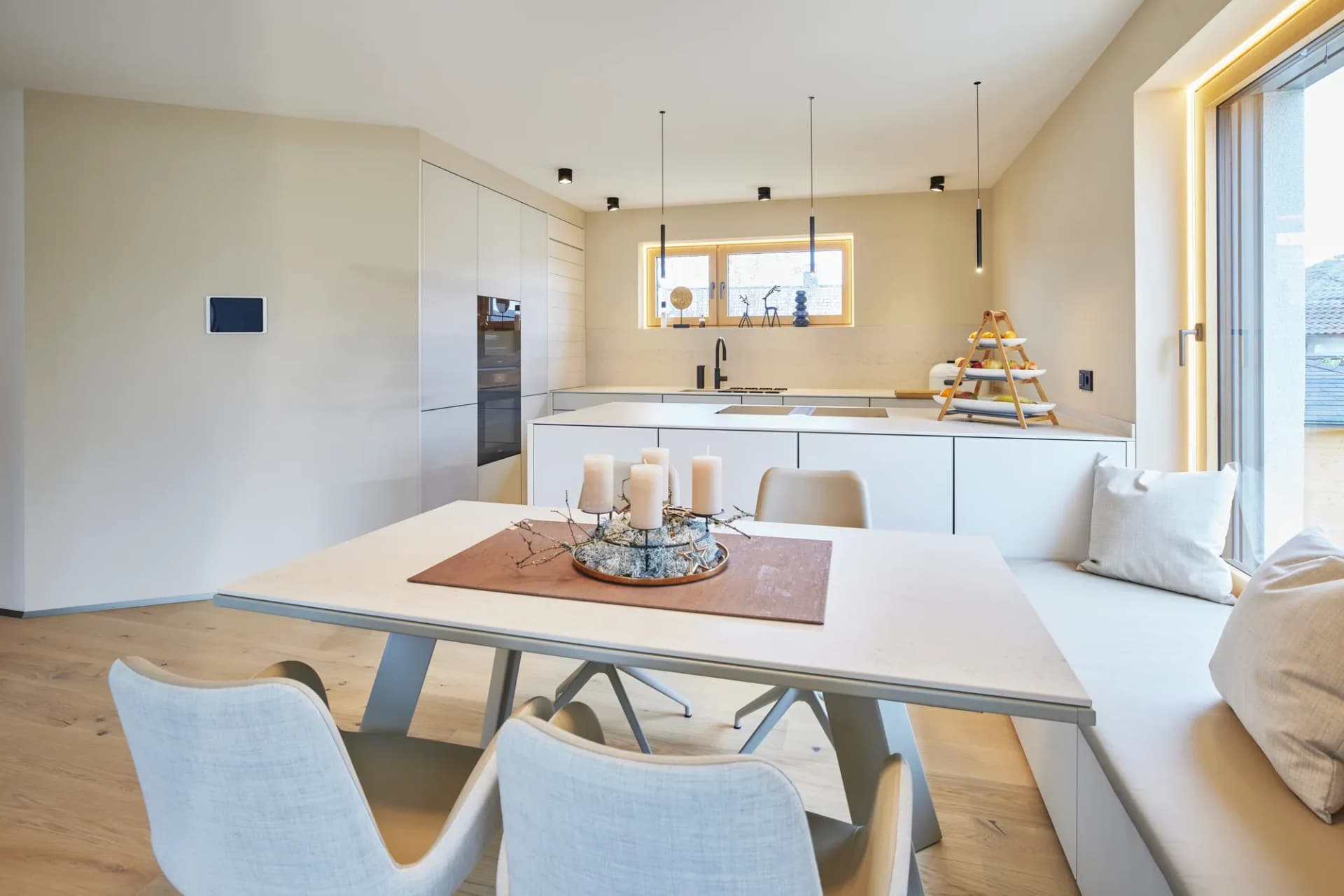 Blick über den gedeckten Esstisch mit Kerzen zur weißen Küchenzeile; warme sandfarbene Wände, Eichenboden und bodentiefes Fenster mit Sitzbank.