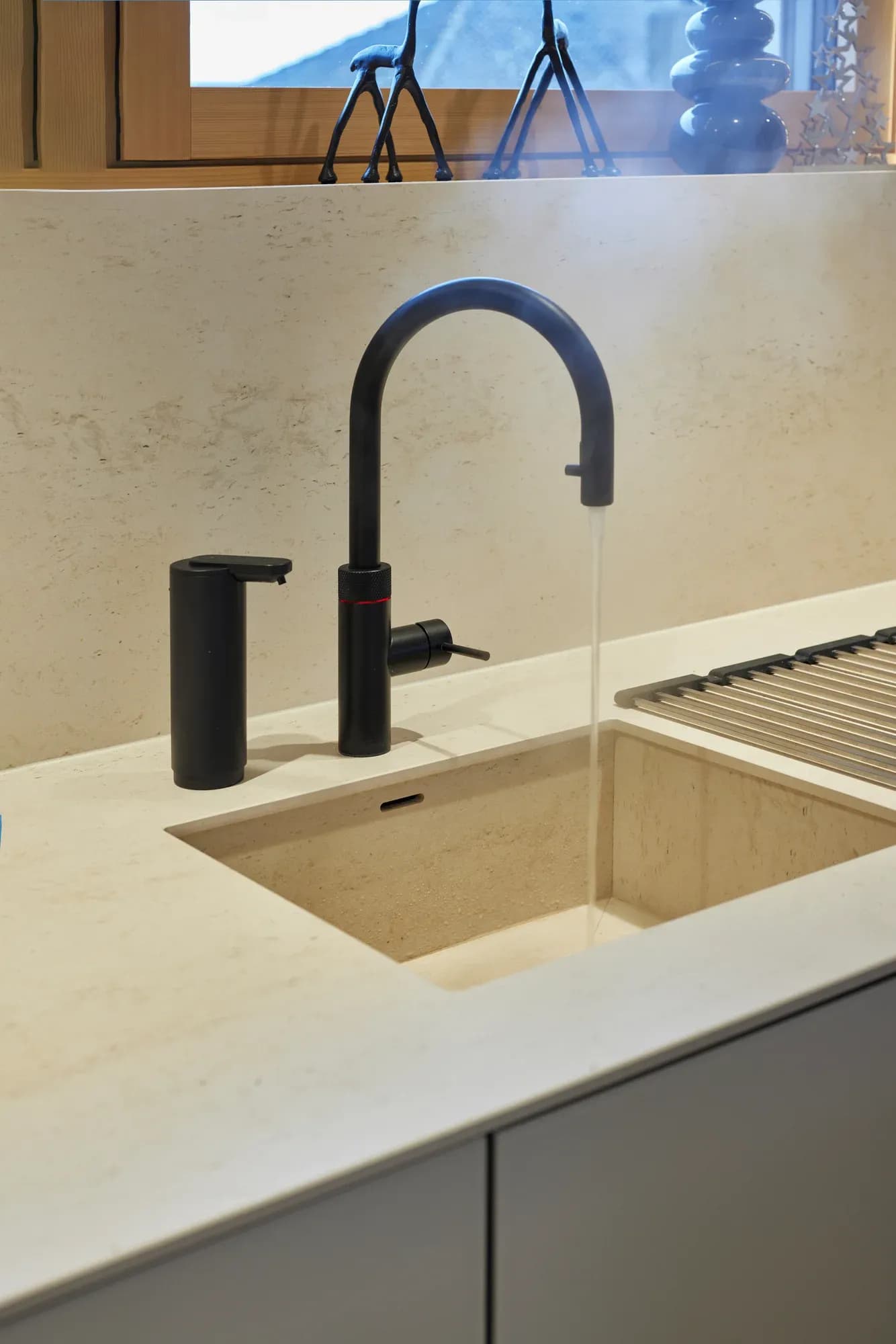 Aus der Natursteinplatte gearbeitetes Unterbaubecken mit schwarzer Quooker-Armatur und schwarzem Seifenspender vor einer steinernen Rückwand.