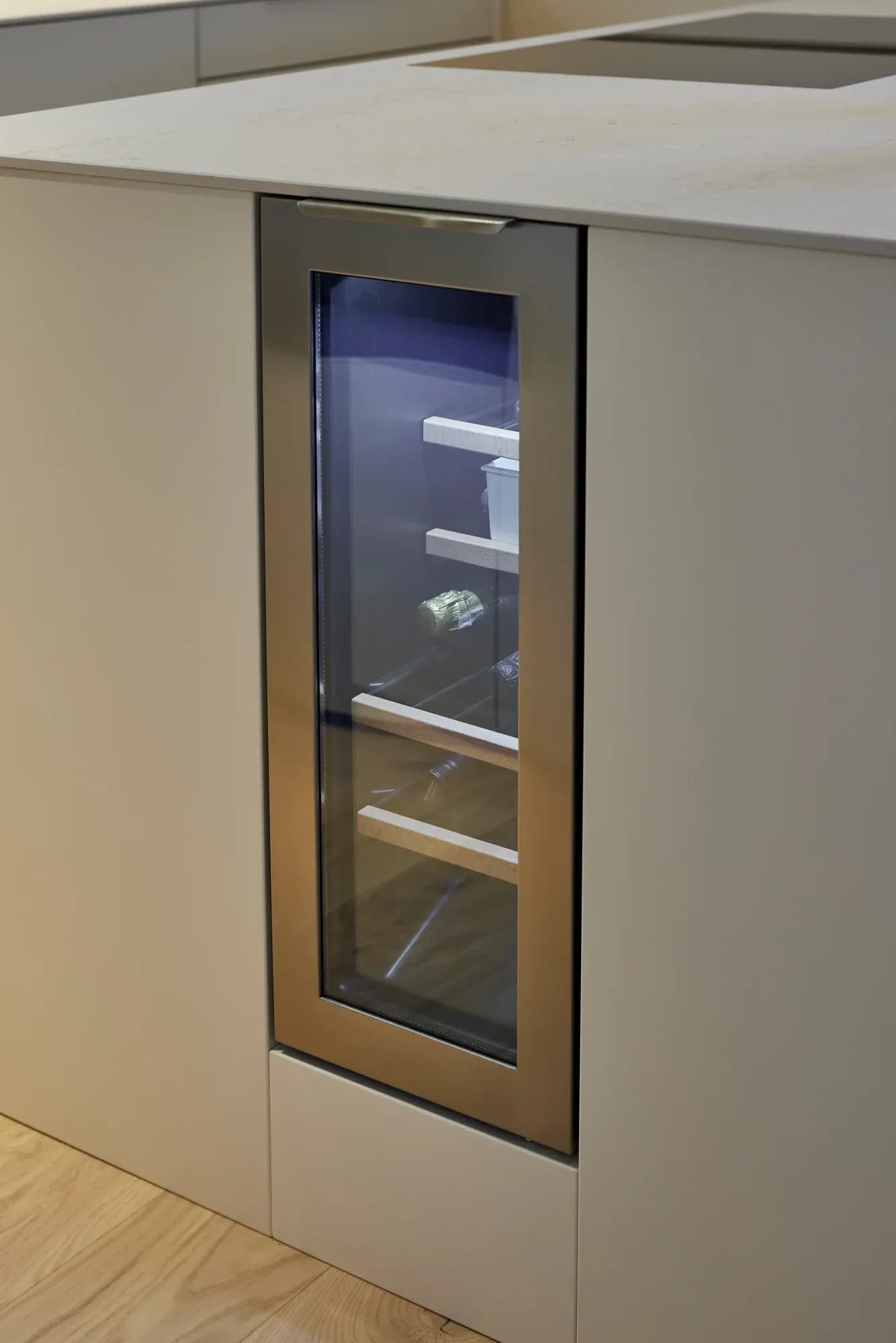 In die Natursteininsel integrierter Weinschrank mit Glastür und Edelstahlrahmen, seitlich in die grifflosen weißen Fronten eingebaut.
