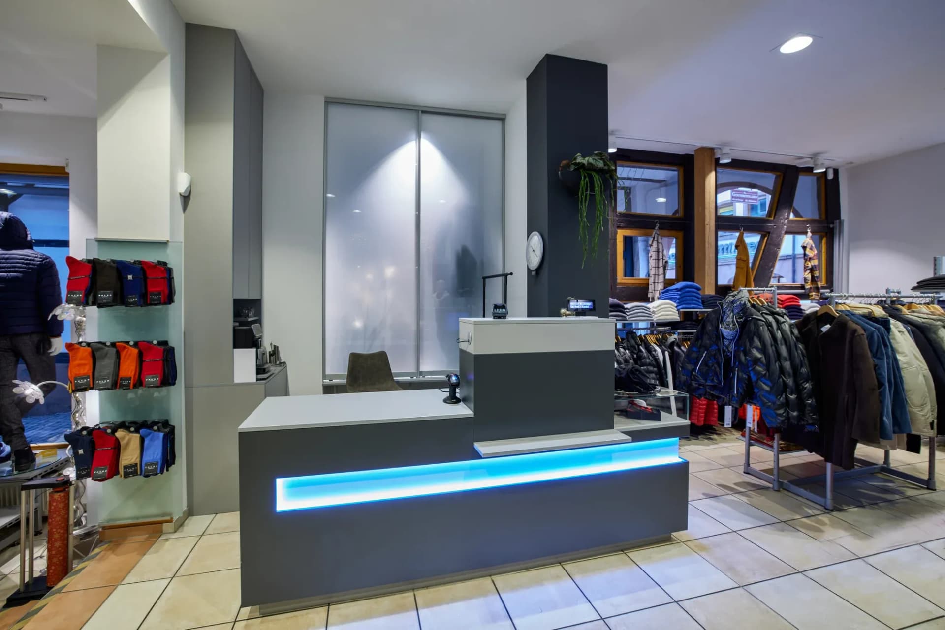 Anthrazitfarbener Verkaufstresen mit heller Arbeitsfläche und blauer LED-Lichtlinie im Herrenmodengeschäft, dahinter Kleiderständer.