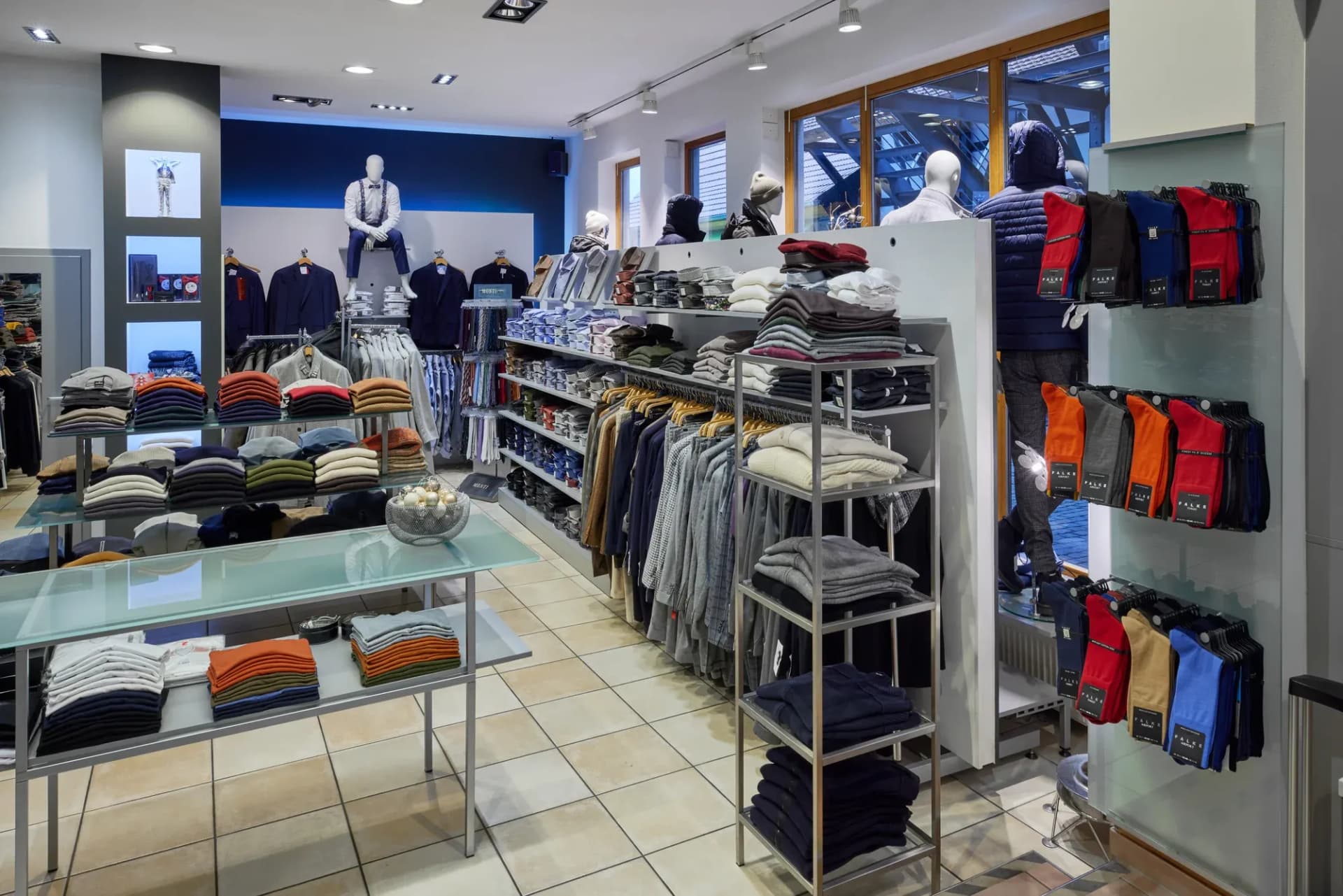Verkaufsraum mit Glastisch, Regalen voller gefalteter Strickware und Kleiderständern.