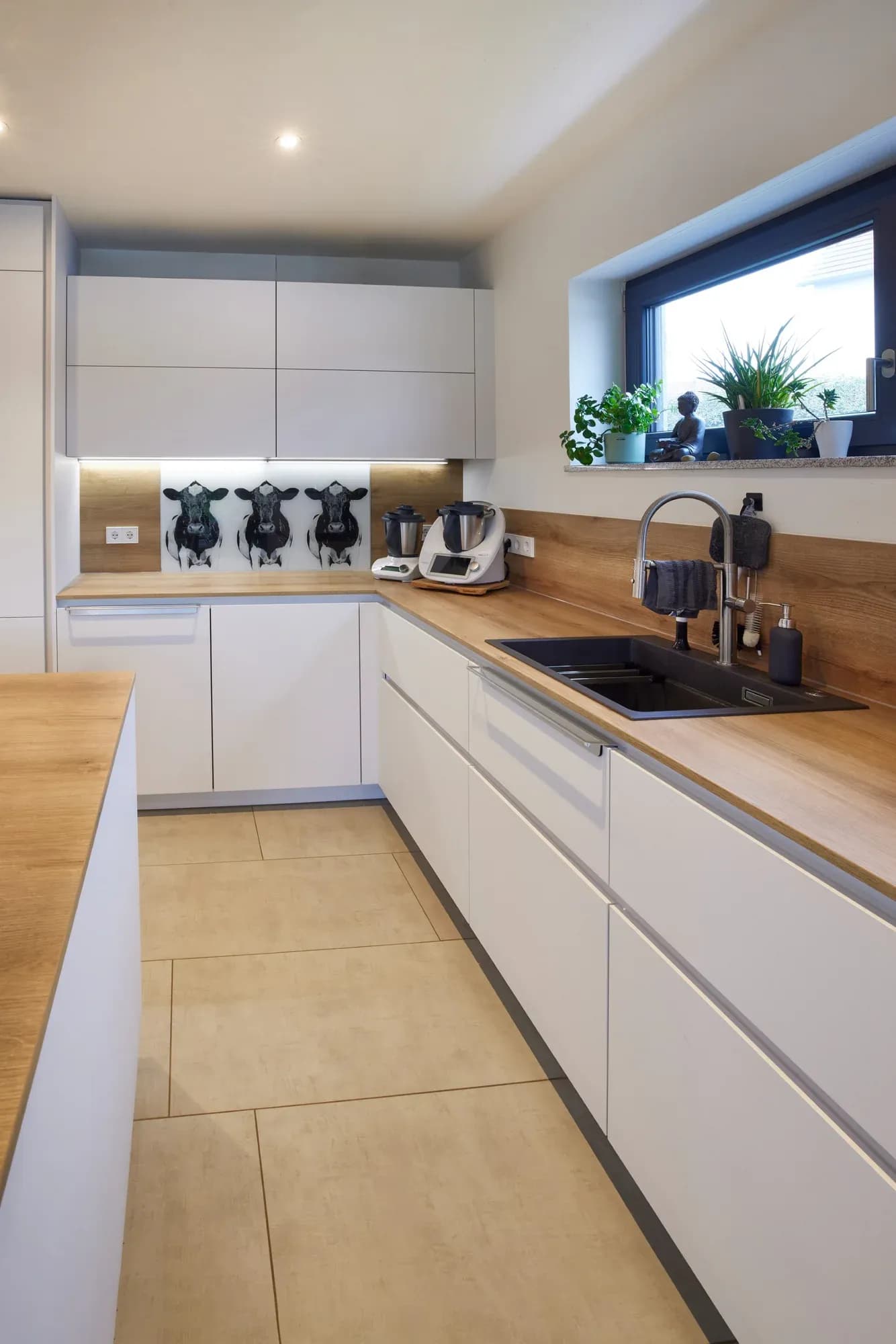 Blick durch die Küche zum Fenster: Zeile mit Backöfen im Hochschrank links, Insel mit Kochfeld rechts, geöffneter Eckschrank mit Karussellauszug.