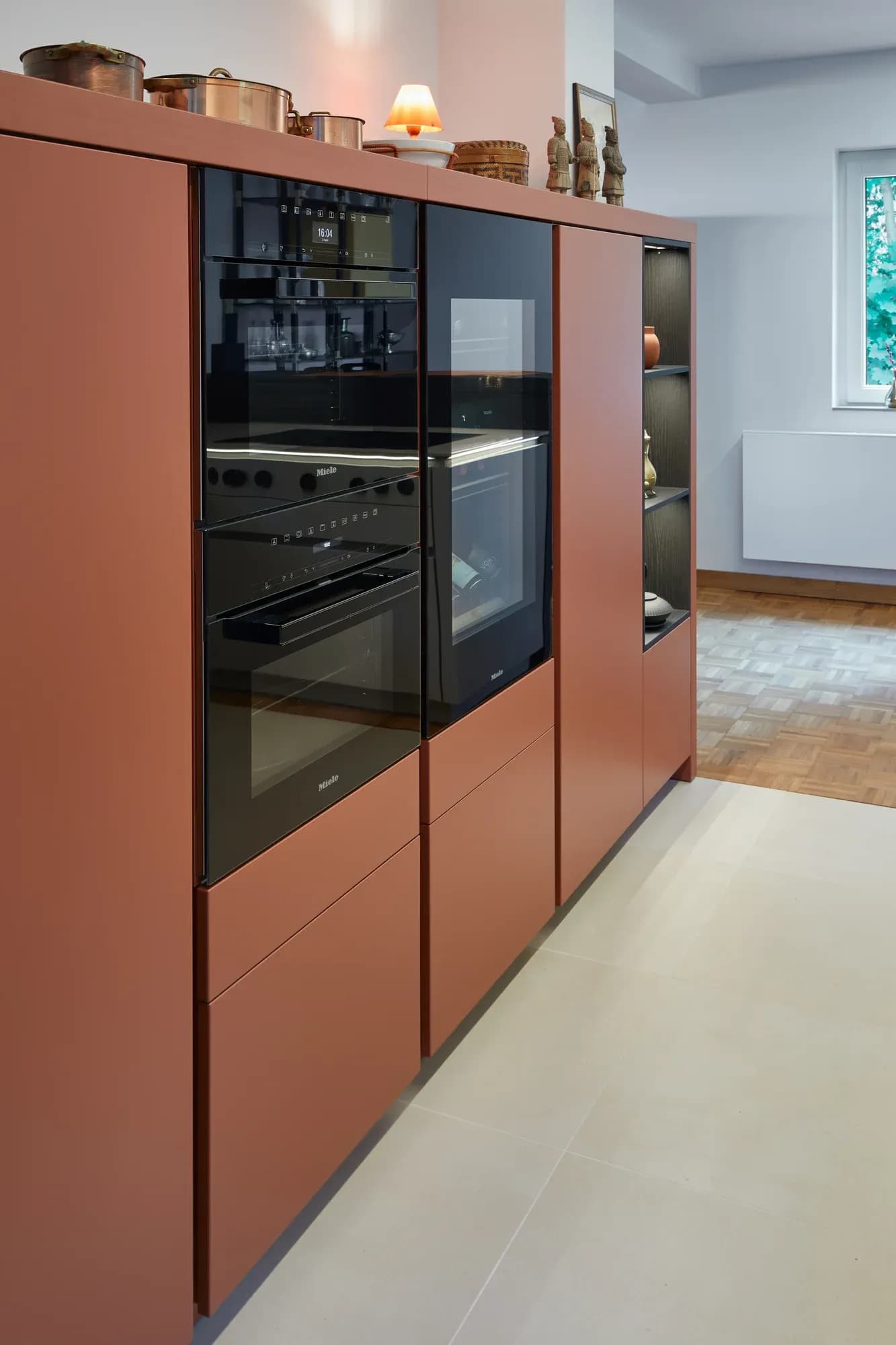 Terrakottafarbener Hochschrank mit eingebauten schwarzen Miele-Backöfen, davor Mosaikparkett und ein Fenster.