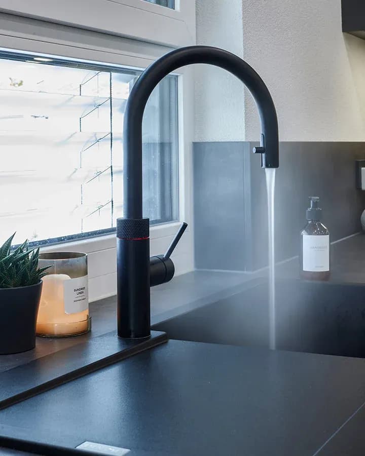 Schwarze Quooker-Armatur mit laufendem Wasser über einem dunklen Spülbecken am Fenster.