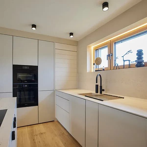 Blick auf die Küchenzeile: hohe Schränke mit schwarzem Miele-Backofen und Dampfgarer, heller Natursteinwerkstoff mit schwarzer Armatur an der Spüle, Fenster mit indirekter Beleuchtung.
