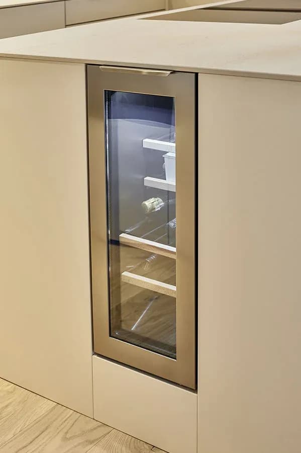 In die Kochinsel integrierter Weinklimaschrank mit Glastür in champagnerfarbenem Rahmen, eingefasst in grifflose Nude-Fronten unter der hellen Natursteinplatte.