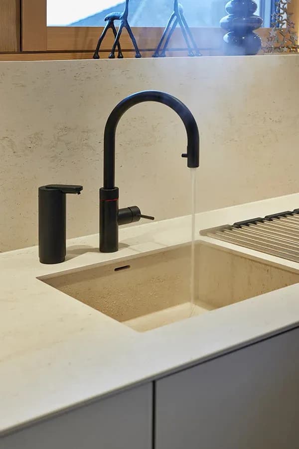 Natursteinspüle mit schwarzem Quooker-Bogenhahn und schwarzem Seifenspender, dahinter die steinerne Rückwand und ein Abtropfrost.