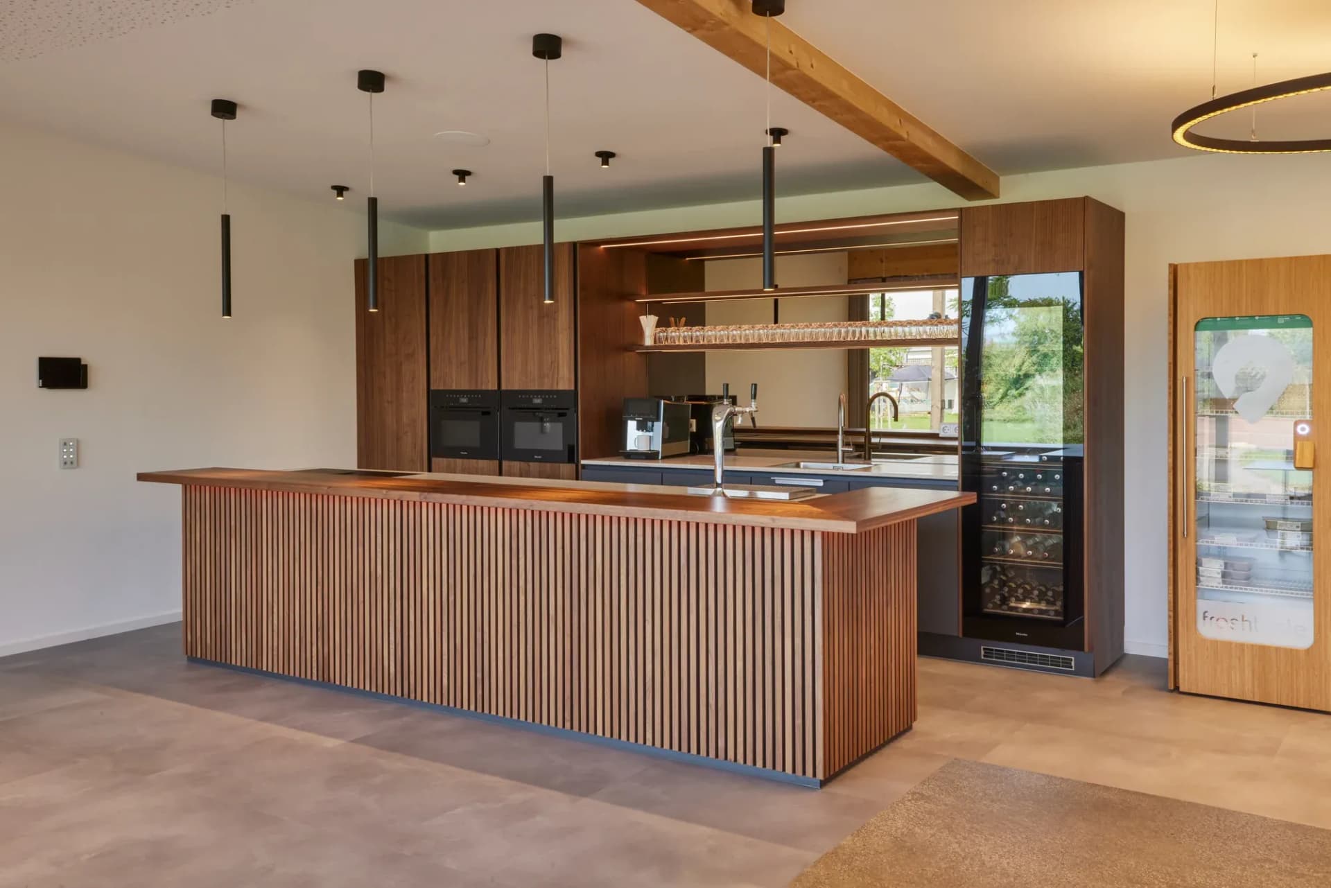 Raumlange Bartheke aus geriffeltem Nussbaum mit massiver Echtholzauflage, dahinter Ausschank, hinterleuchtete Gläserablage und ein Weinklimaschrank; darüber ein sichtbarer Dachbalken aus Holz.