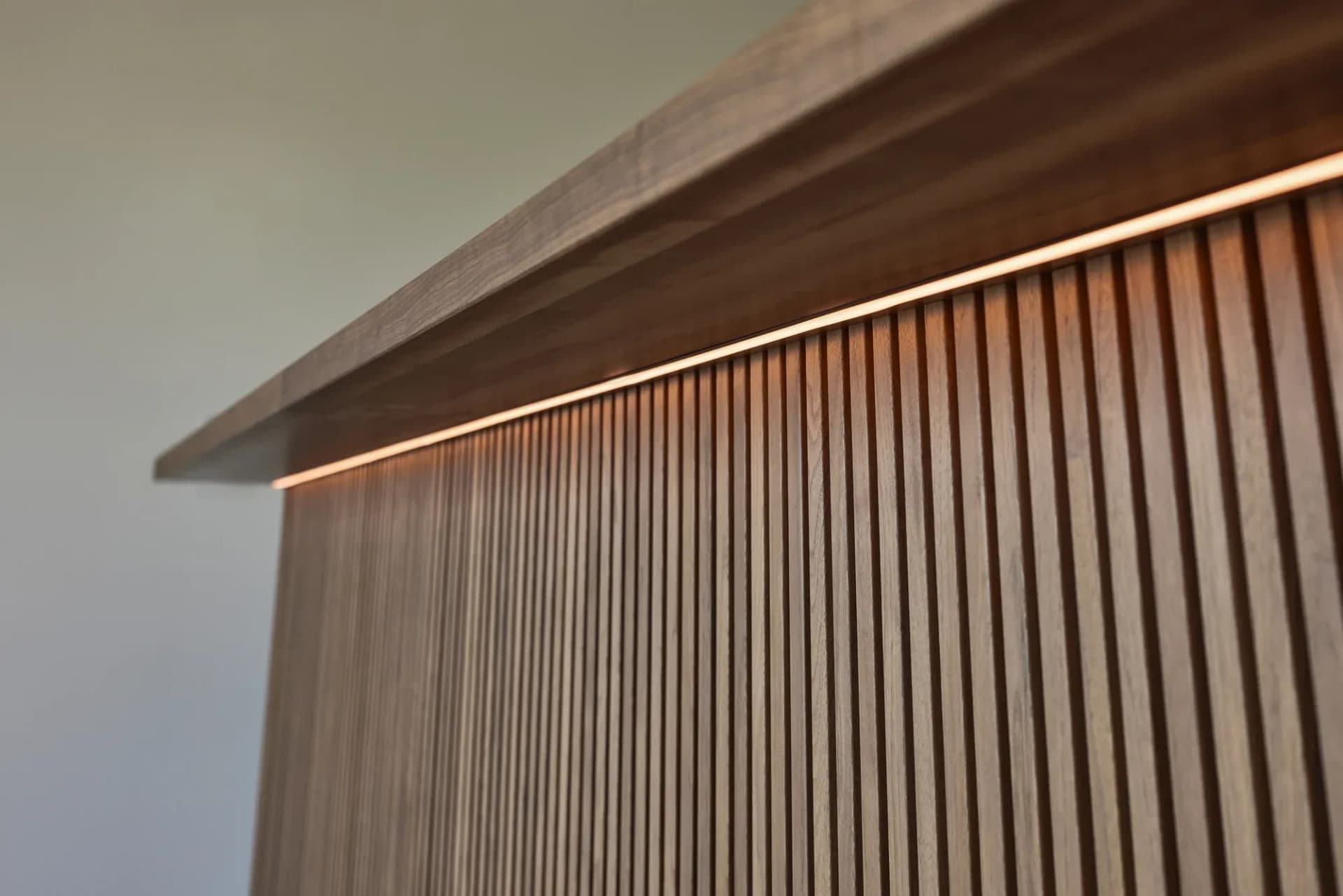 Detail der Theke: massive Nussbaumauflage über der geriffelten Lamellenfront, an der Unterkante eine durchlaufende warme Lichtlinie.