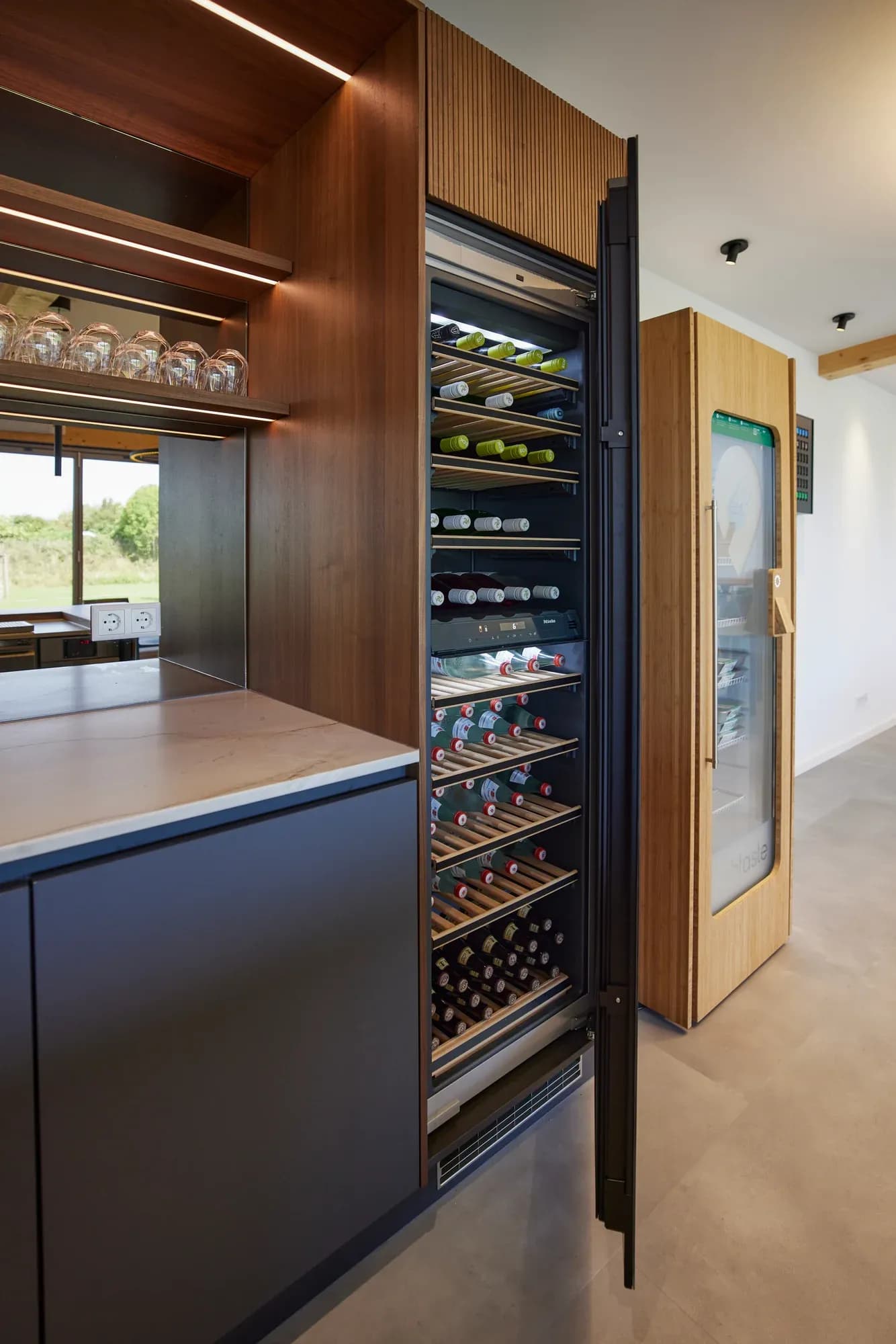 Geöffneter Weinklimaschrank hinter Glas, gefüllt mit Wein- und Getränkeflaschen auf Holzauszügen; rechts ein gekühlter Selbstbedienungsschrank.