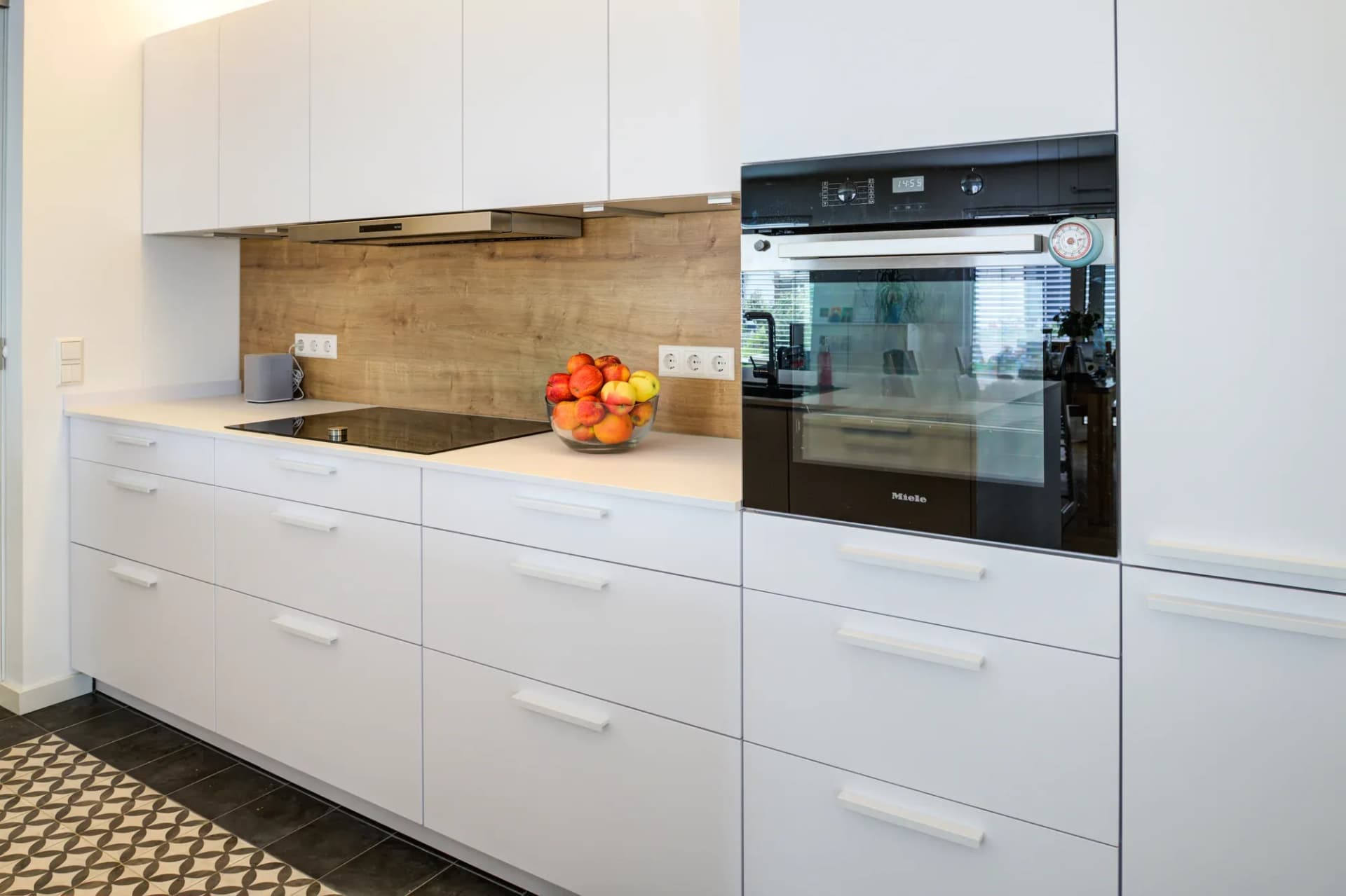 Küchenzeile mit Induktionskochfeld, Holzoptik-Rückwand und Miele-Backofen in der weißen Fronten-Wand, davor der Boden aus gemusterten Zementfliesen.