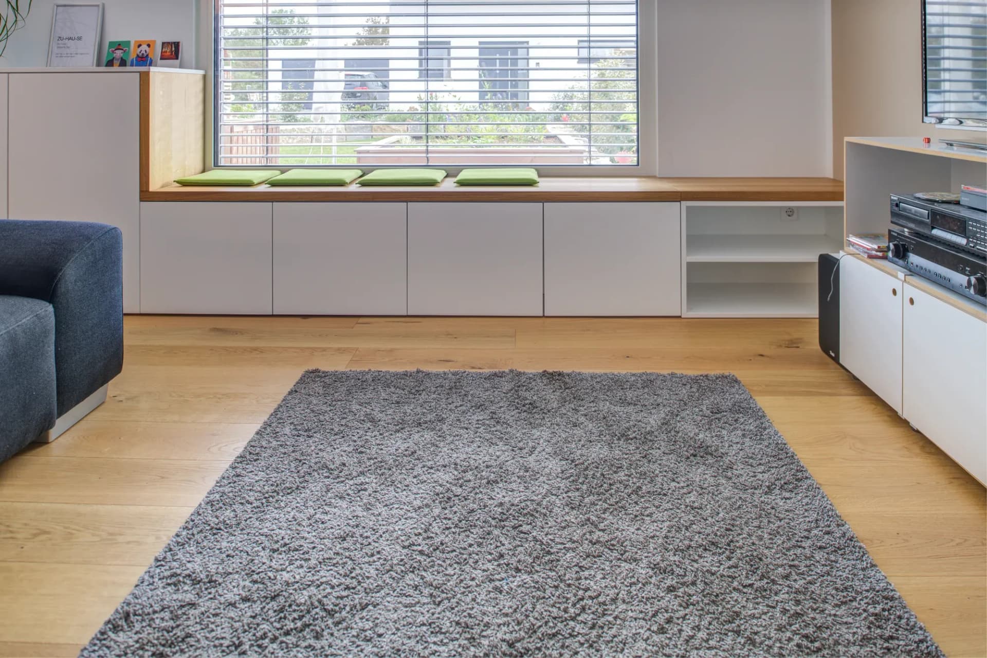 Wohnzimmer mit langem weißem Einbau-Lowboard, Eichenauflage als Fensterbank mit grünen Sitzkissen und offenem Regalfach, davor ein grauer Teppich.
