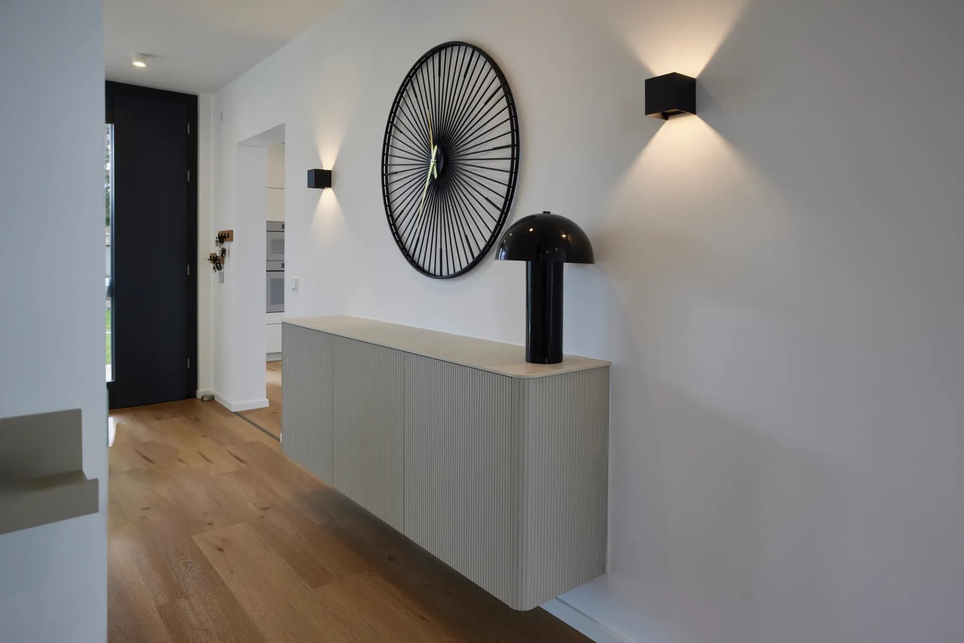 Sideboard mit Rillenfront im Eingangsbereich, darüber ein großes Rad als Wandobjekt und schwarze Wandleuchten.