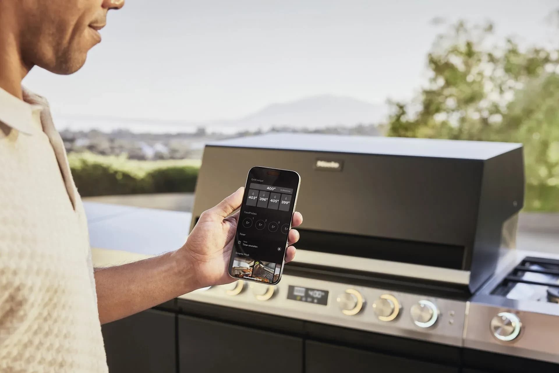 Mann kontrolliert auf der Terrasse per Smartphone-App Temperaturen und Timer seines Miele Outdoor-Grills, im Hintergrund Landschaft und Berge