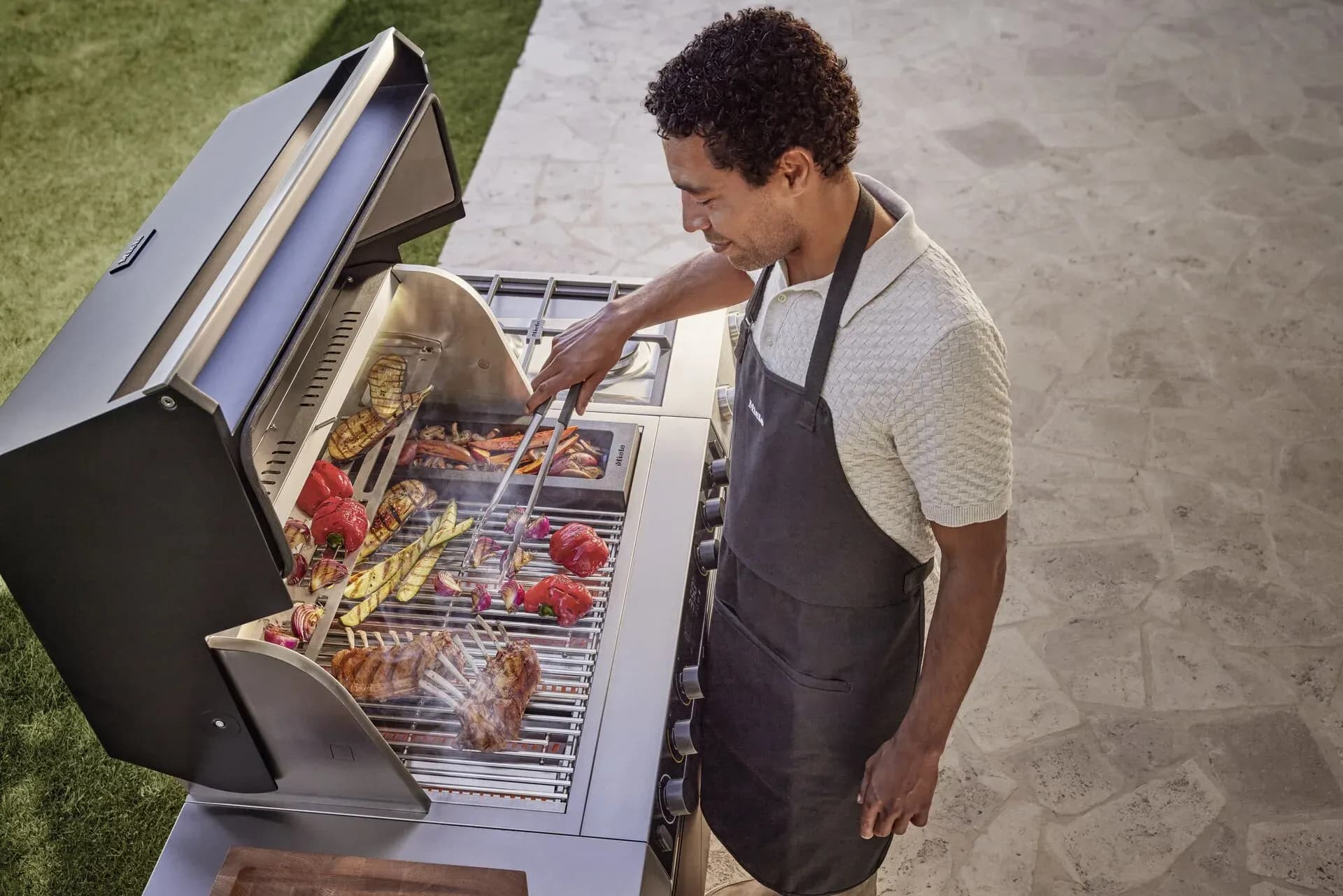 Mann mit Schürze grillt Lammkoteletts und Gemüsespieße auf einem geöffneten Miele-Grill