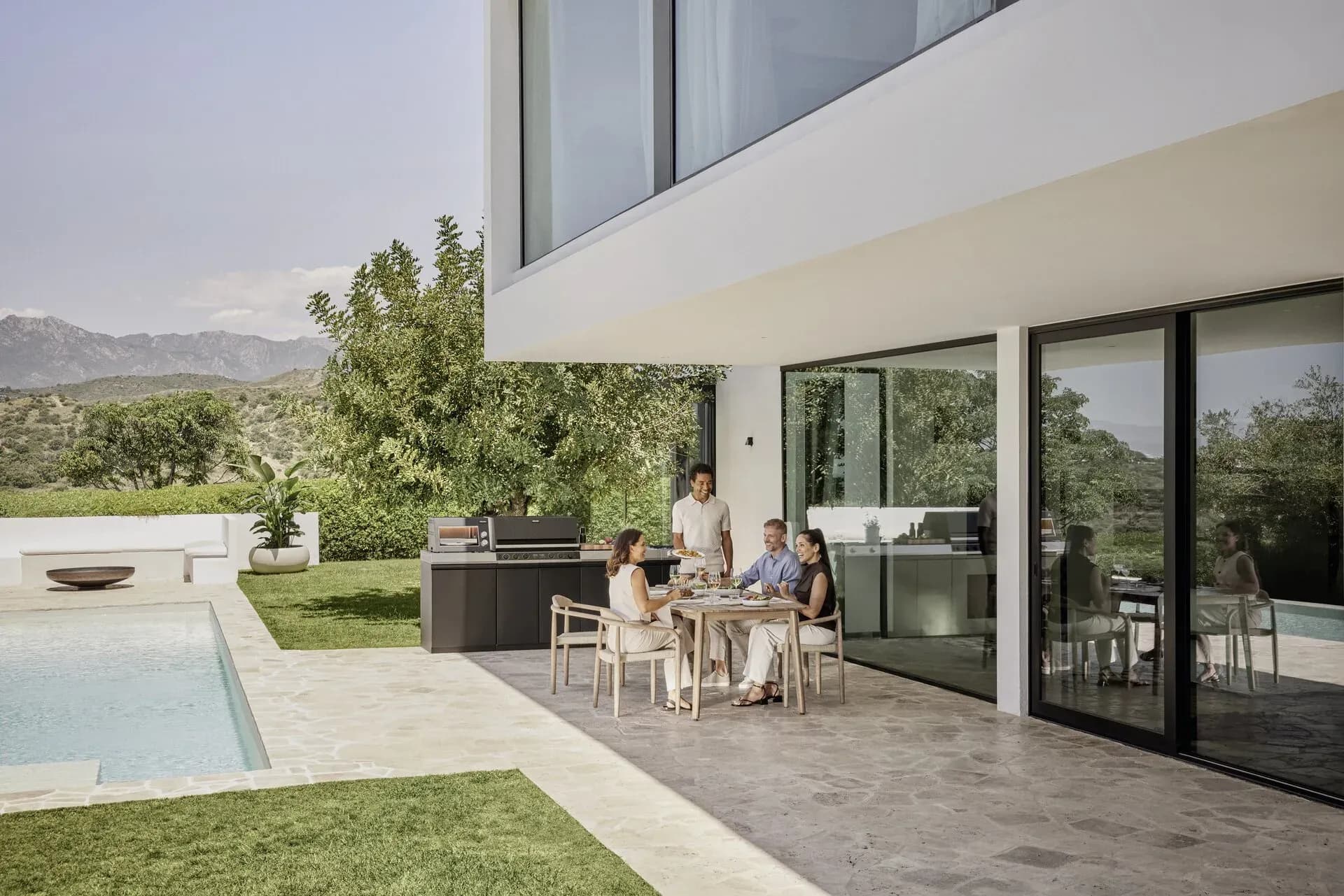 Vier Freunde sitzen lachend an einem Tisch auf der Terrasse einer modernen Villa mit Pool, im Hintergrund eine dunkle Miele Outdoor-Küche und Berge