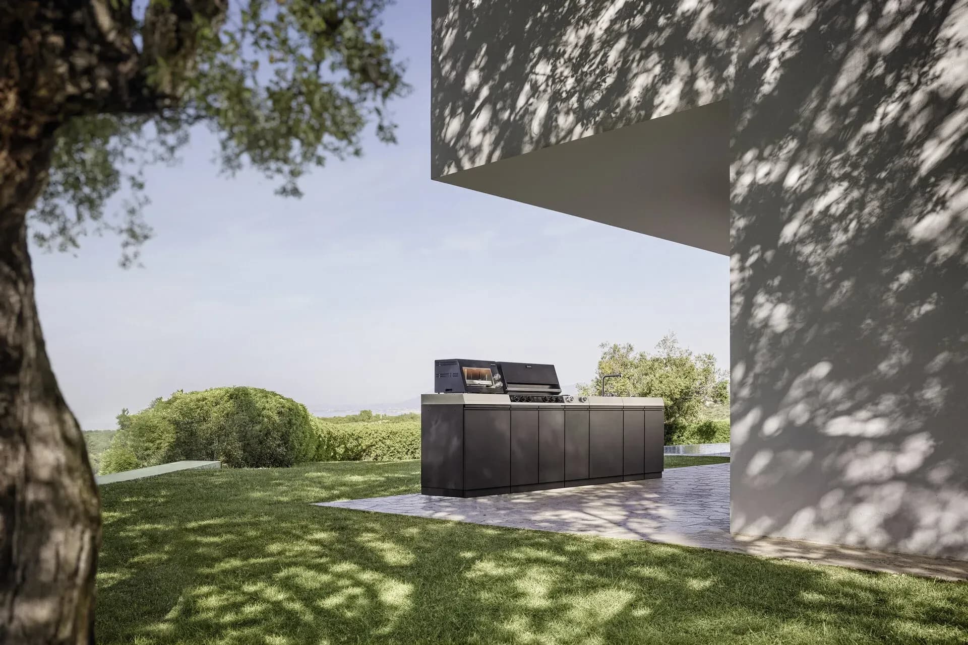 Freistehende, anthrazitfarbene Miele Outdoor-Küche auf einer Natursteinterrasse unter einem Dachüberstand, dahinter Rasen, ein alter Baum und Hügellandschaft