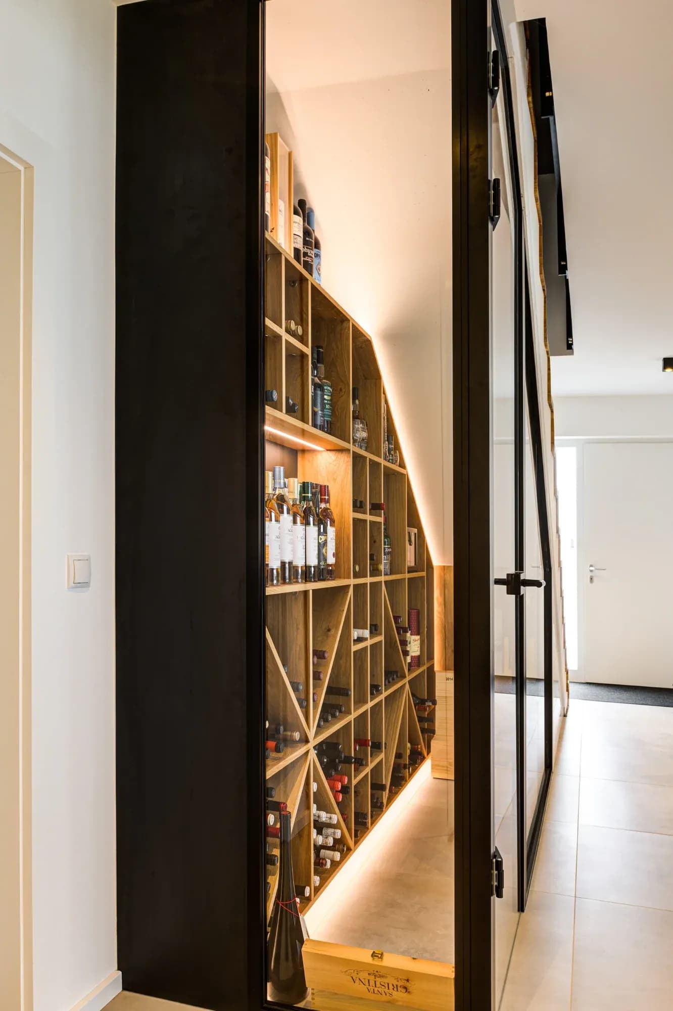 Geöffnete Stahl-Glas-Tür gibt den Blick in den Weinkeller frei: ein warm beleuchtetes Eichenregal folgt der Treppenschräge, gefüllt mit Wein- und Whiskyflaschen.