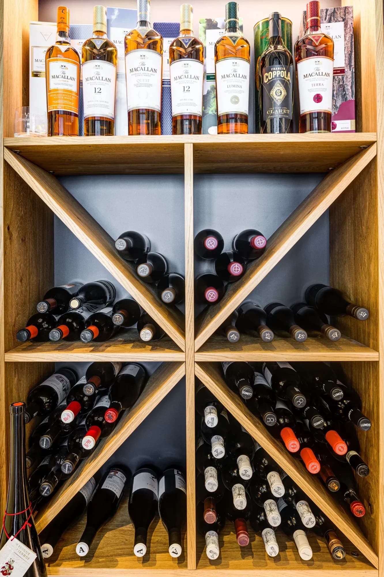 Symmetrisches Kreuzregal aus Eiche, dicht mit liegenden Weinflaschen belegt, darüber eine Reihe stehender Whiskyflaschen.