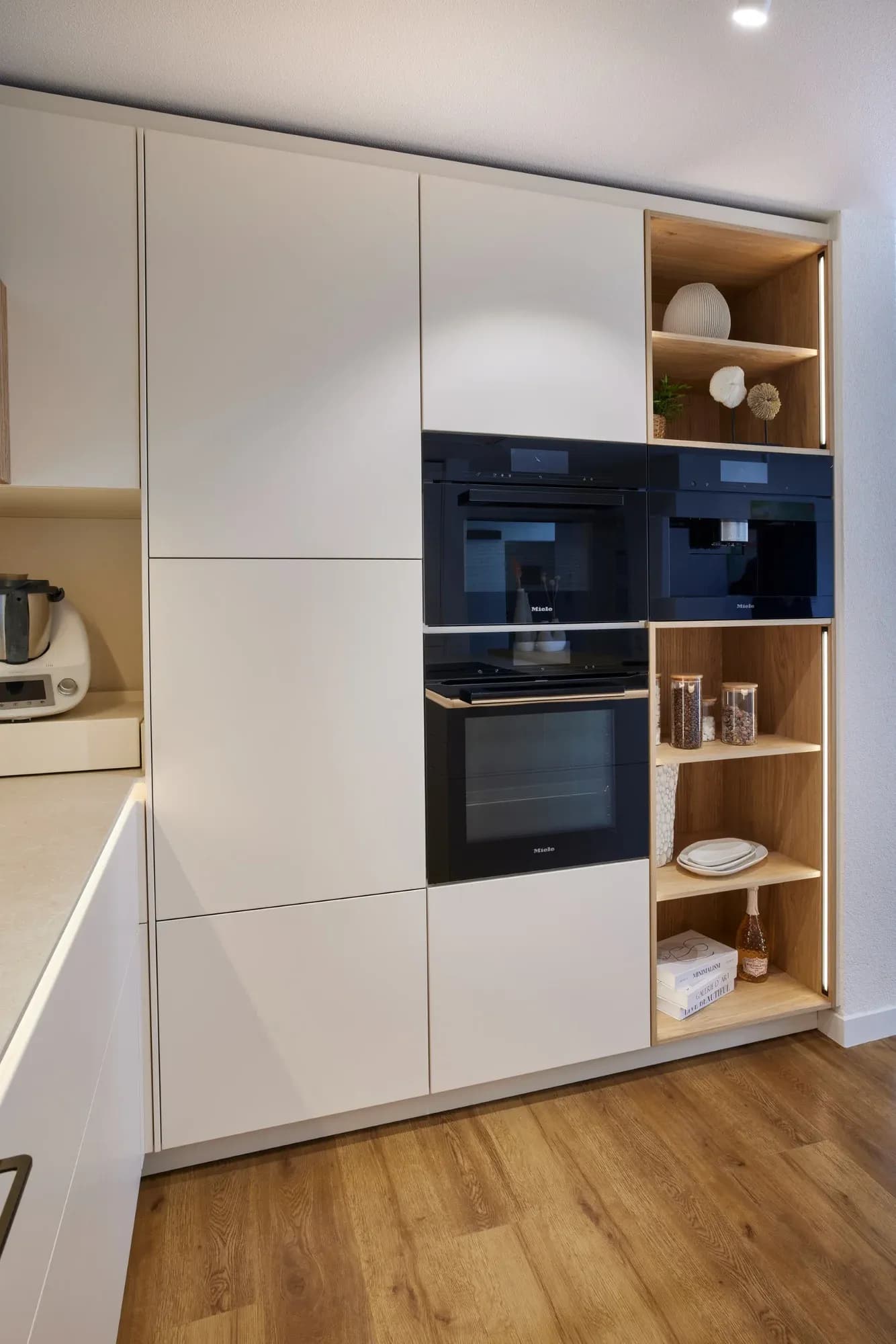 Hochschrankwand in Sandbeige mit gestapelten Miele-Geräten in Obsidianschwarz (Backofen, Dampfgarer, Kaffeeautomat) und einem offenen Eichen-Regal mit indirekter Beleuchtung.