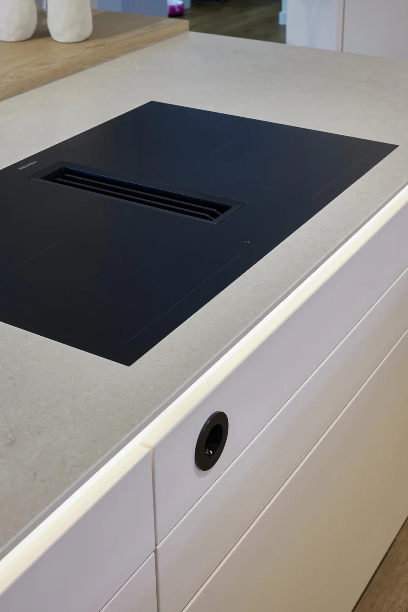 Detail des mattschwarzen Miele-Induktionskochfelds mit integriertem Abzug in der hellen Keramik-Arbeitsplatte, darunter ein warmer Lichtsaum an der Inselkante.