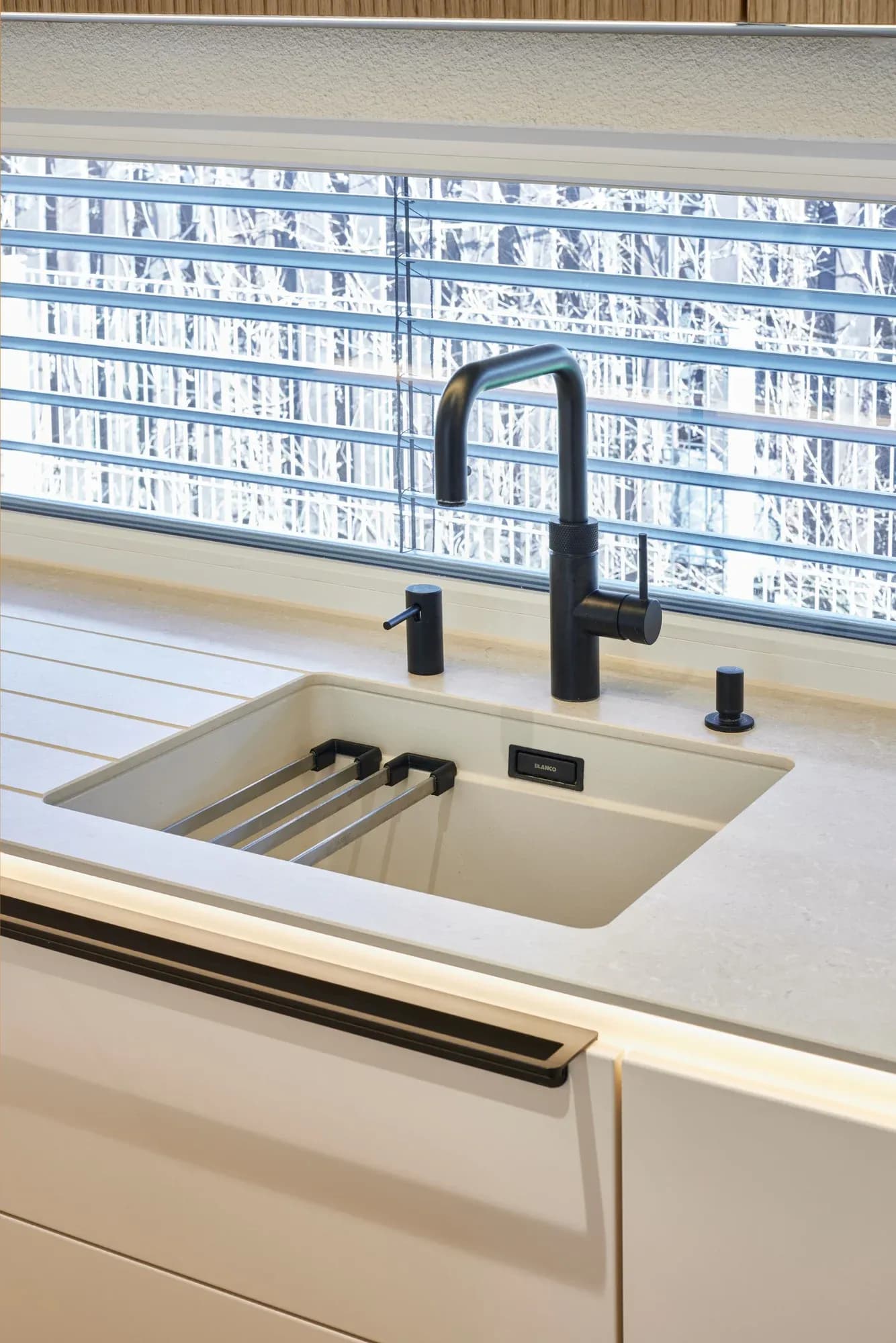 Spülplatz am Fenster: Blanco-Unterbaubecken in Softweiß mit mattschwarzer Quooker-Armatur und Seifenspender, helle Keramik-Arbeitsplatte mit warmem Lichtsaum.