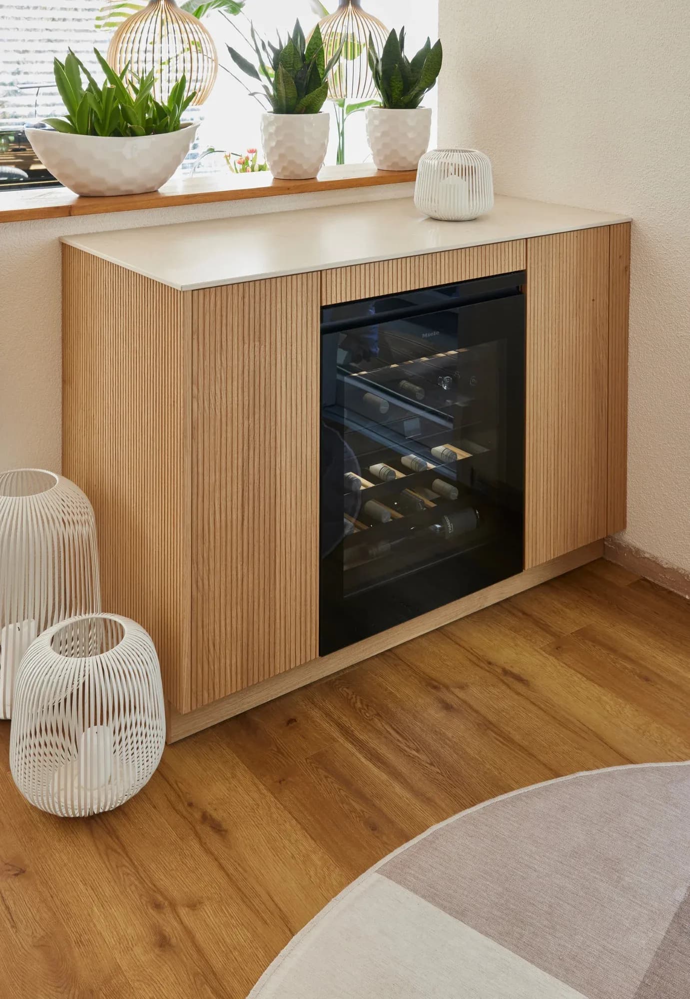 Sideboard aus geriffelter Eiche mit heller Steinplatte, darin ein eingebauter Miele-Weinschrank hinter Glas, davor Bodenleuchten und Zimmerpflanzen am Fenster.